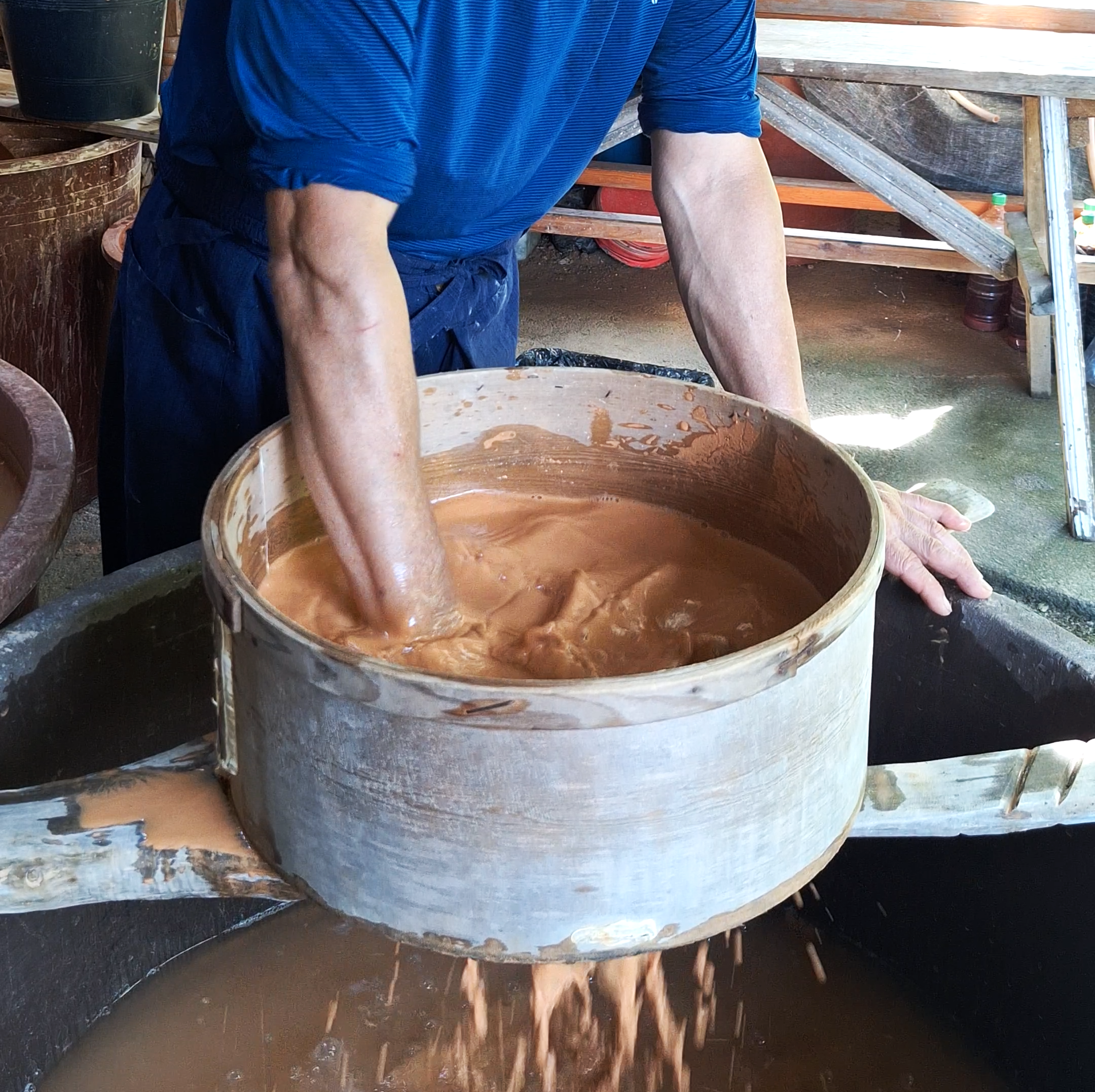 A person in a blue shirt stirring a large wooden tub filled with a brown liquid, possibly chocolate, with a spoon or paddle, in an outdoor workshop or farm setting.