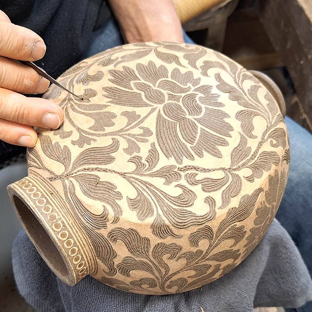A person working on decorating a ceramic vase with intricate floral patterns using a fine brush or tool.