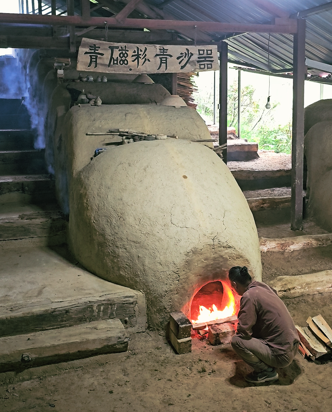 A person kneeling in front of a traditional clay kiln with a glowing fire inside, used for pottery or ceramic making, under a roofed structure with a signboard with Chinese characters.