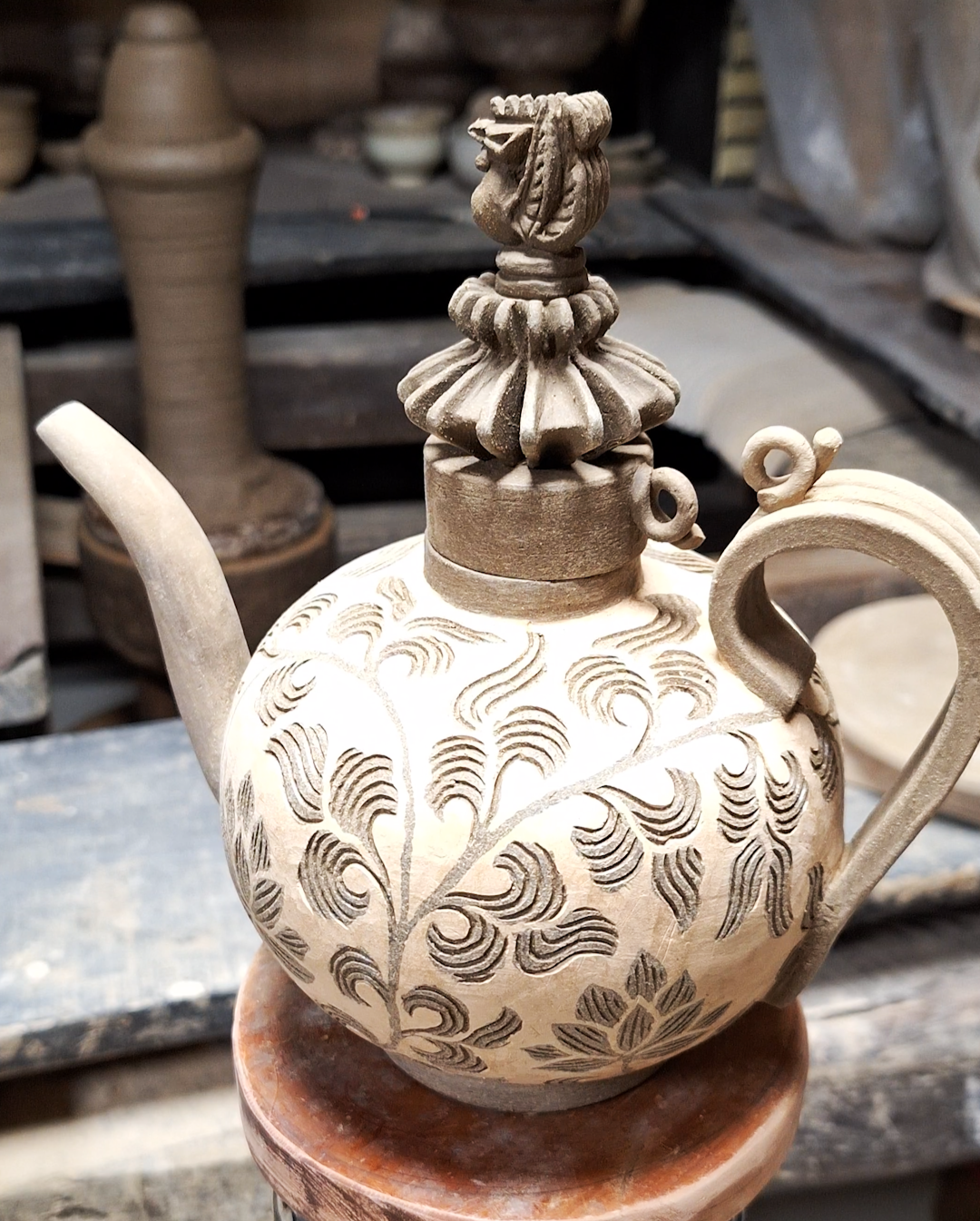 A decorative ceramic teapot with intricate carved floral and leaf patterns, sitting on a wooden stand in a pottery workshop background.