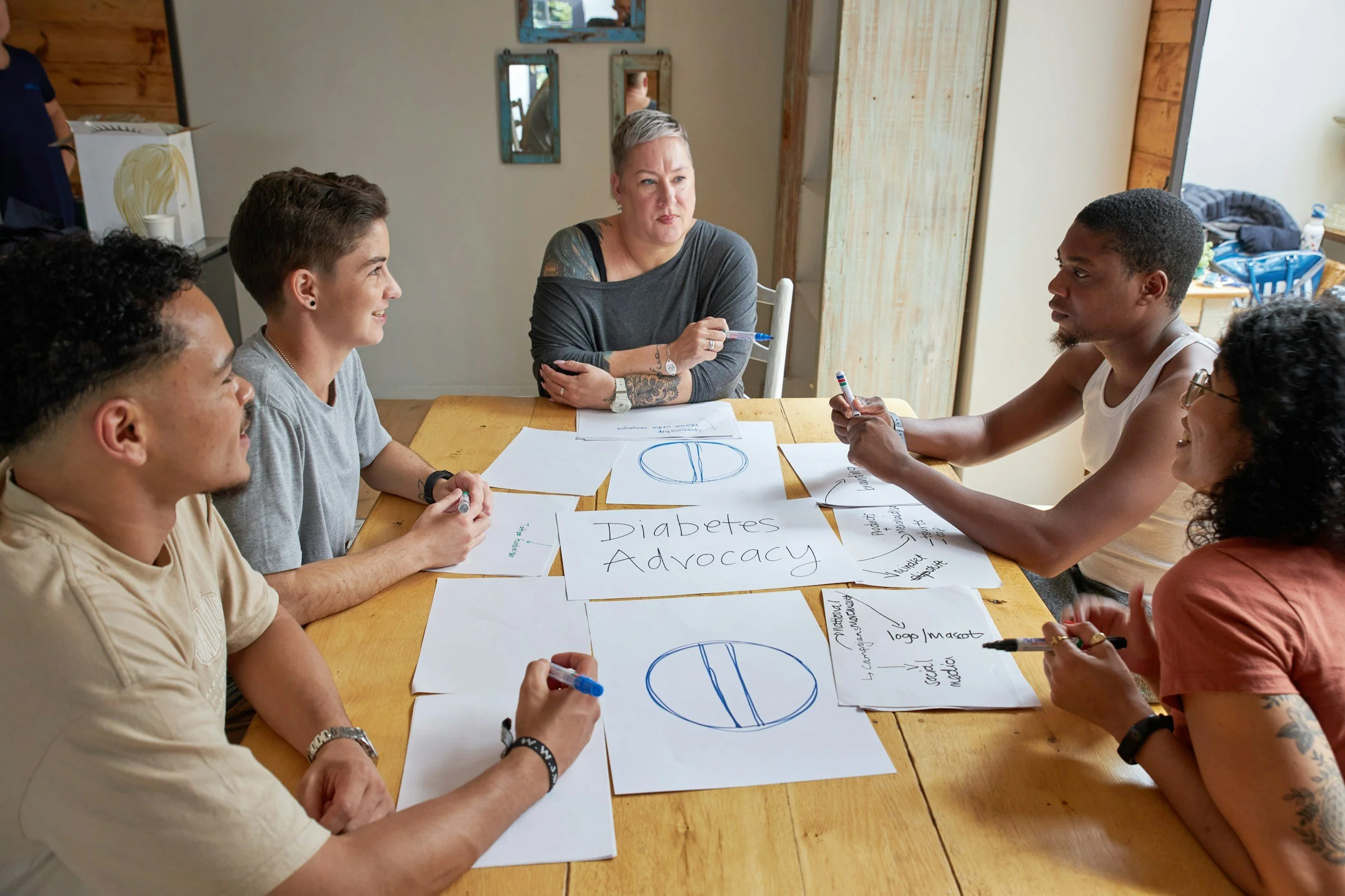 A diverse group of six people sitting around a wooden table having a discussion about diabetes advocacy, with notes and diagrams on paper in front of them.