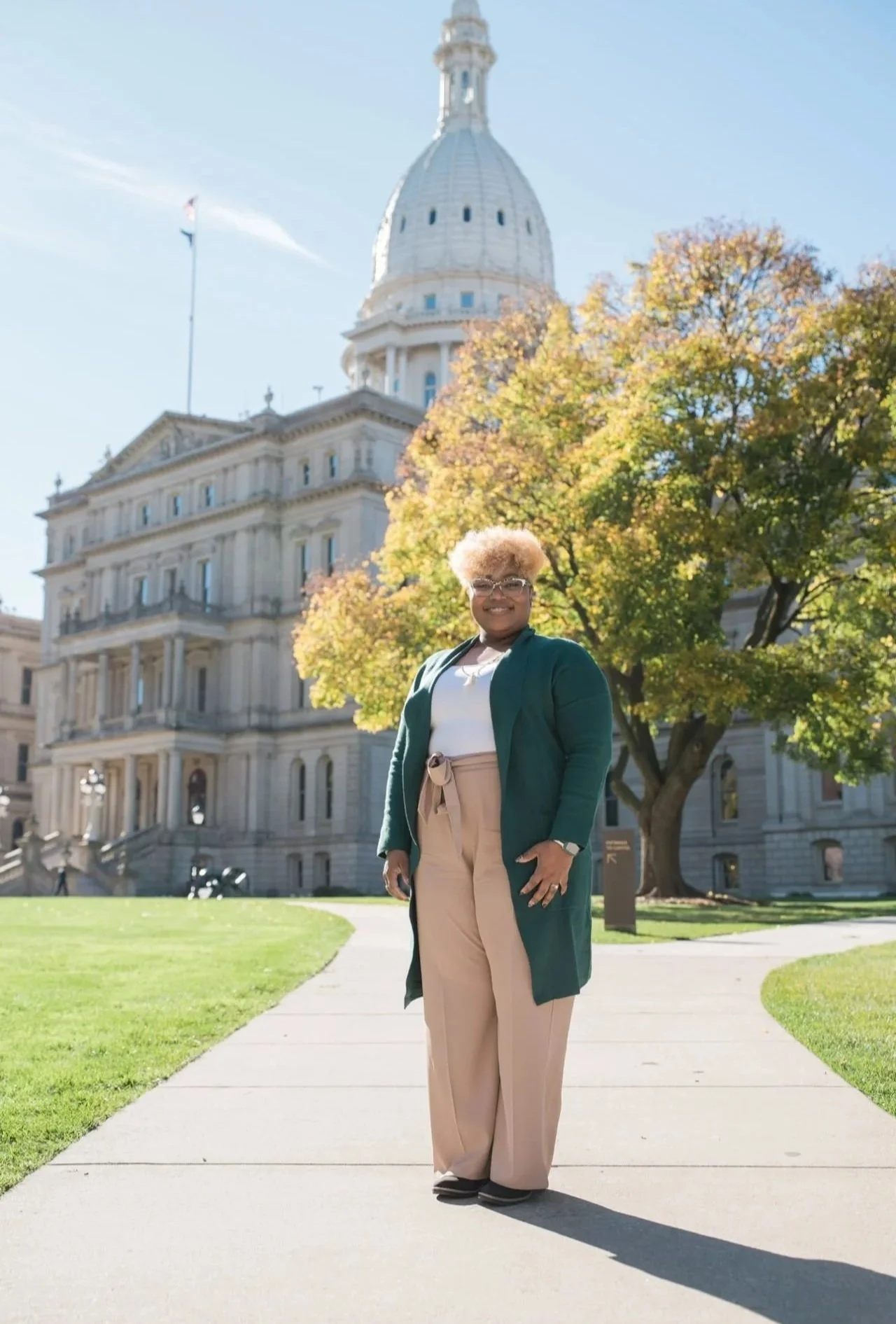 A woman with short blonde hair and glasses standing on a sidewalk in front of a historic government building with a large dome, surrounded by autumn trees and green grass.
