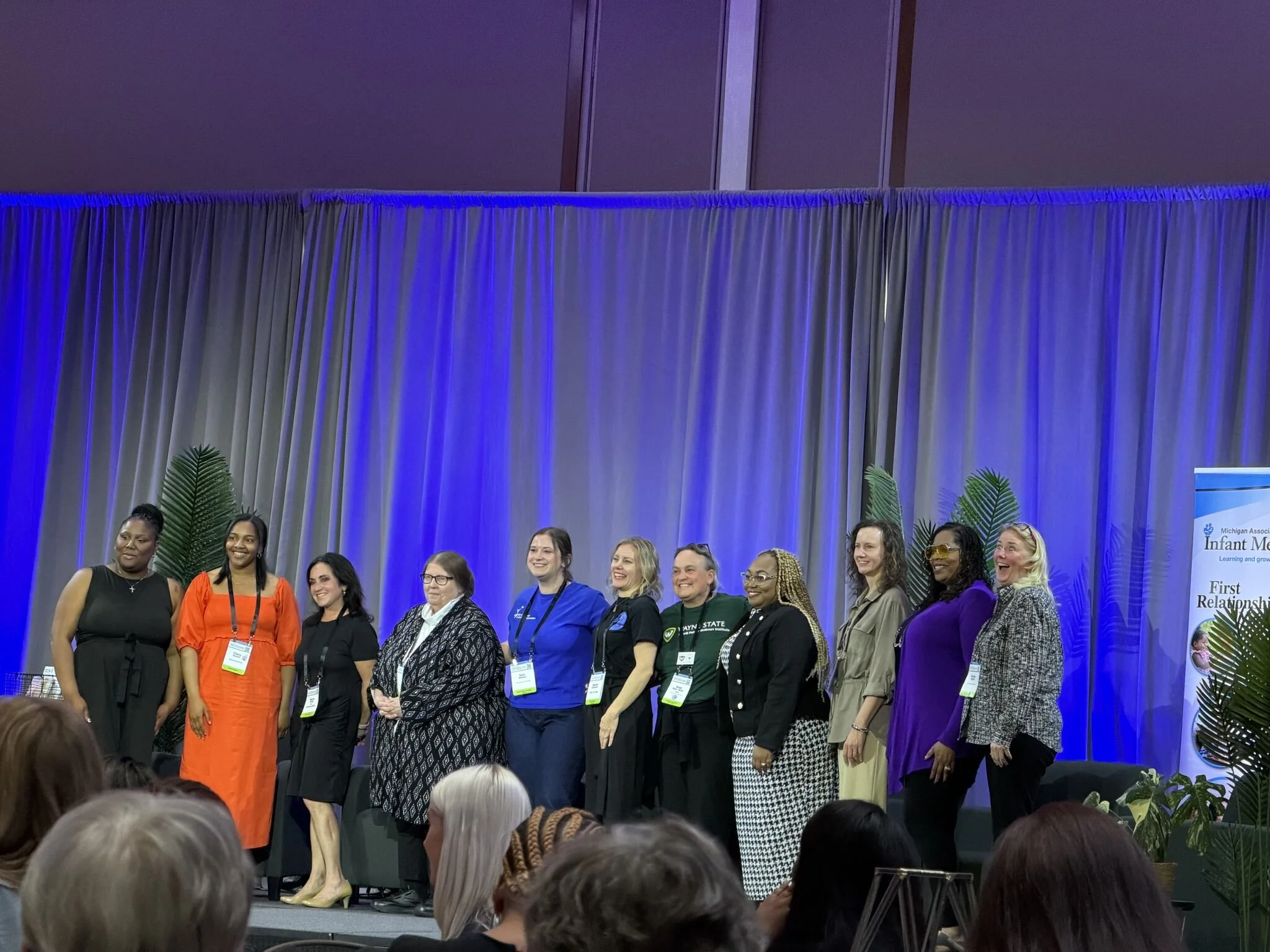 A group of diverse women standing on a stage at a conference, smiling. They are wearing conference badges and are positioned in front of a blue-lit curtain and a sign that reads 'Michigan Association for Infant Mental Health.'