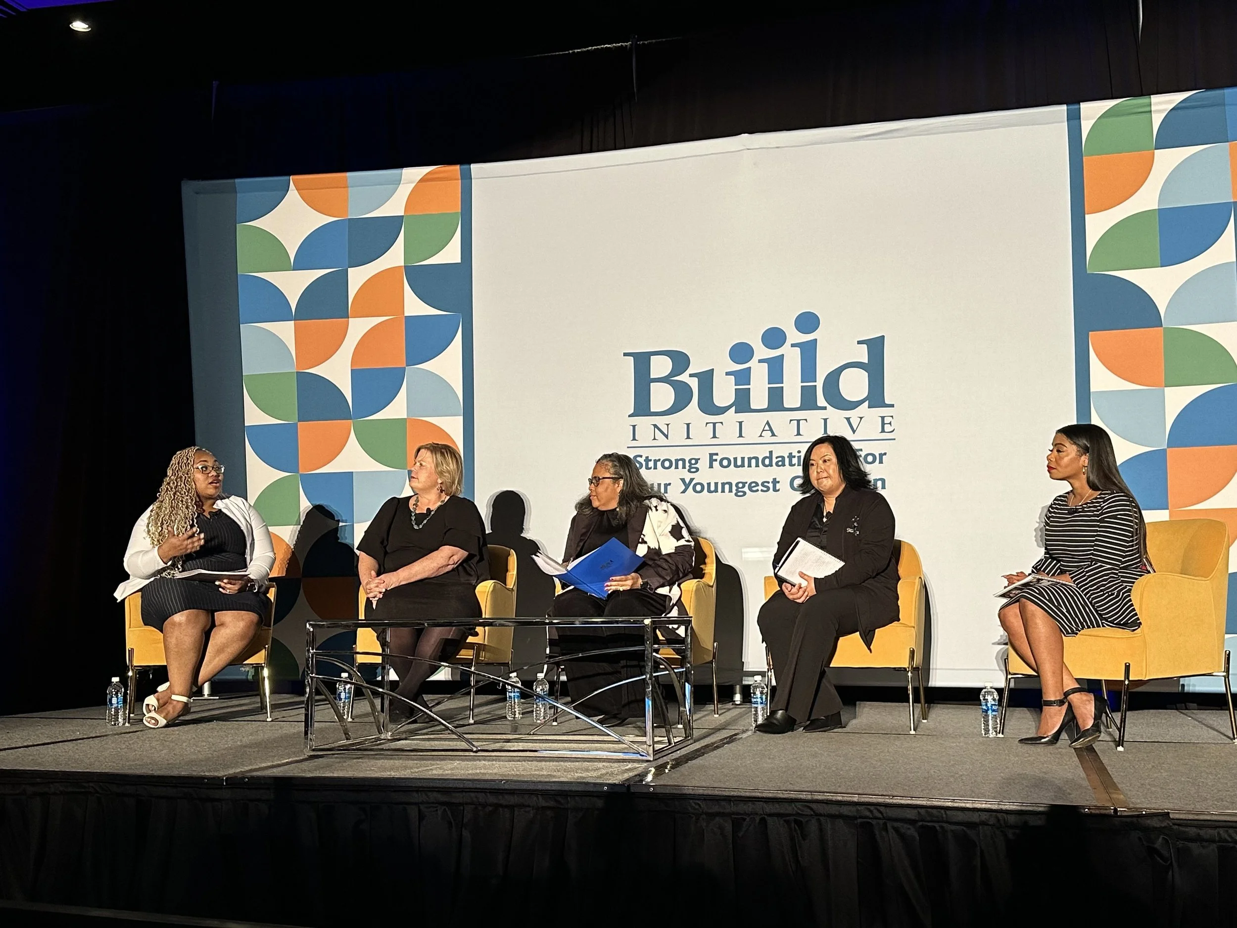 A group of six women seated on stage in front of a large banner that reads 'Build Initiative' and 'Strong Foundation for Our Youngest Children.' The women are engaged in discussion, some holding microphones and papers. The stage has yellow chairs and water bottles placed on the floor. The background features a decorative geometric pattern with blue, green, orange, and white shapes.