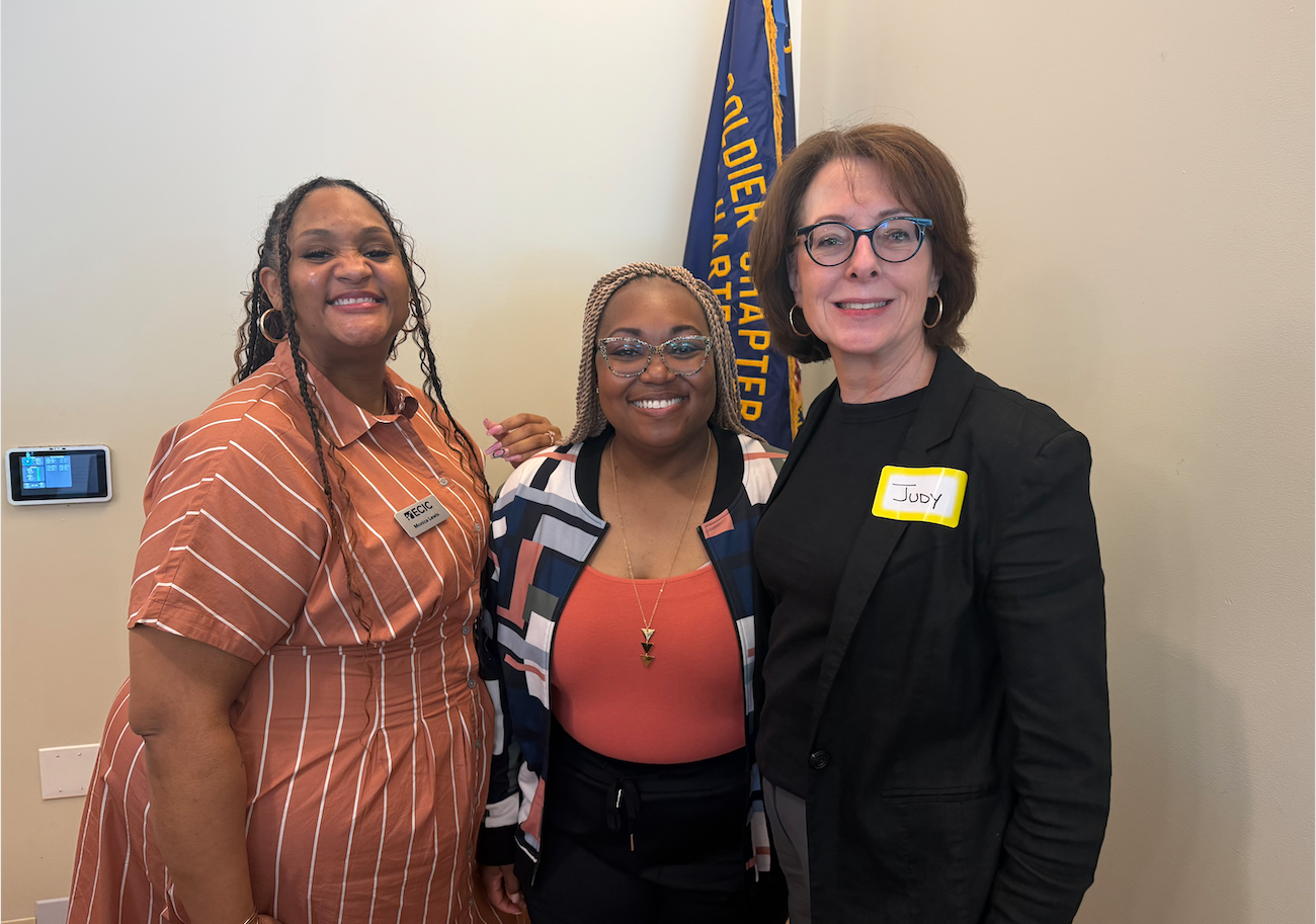 Three women standing together and smiling at the camera in an indoor setting, with a flag in the background. The woman on the left is wearing a rust-colored striped dress and a name tag. The woman in the middle is wearing glasses, a patterned jacket, and an orange top. The woman on the right is wearing glasses, a black jacket, and a yellow name tag with the name "Judy."
