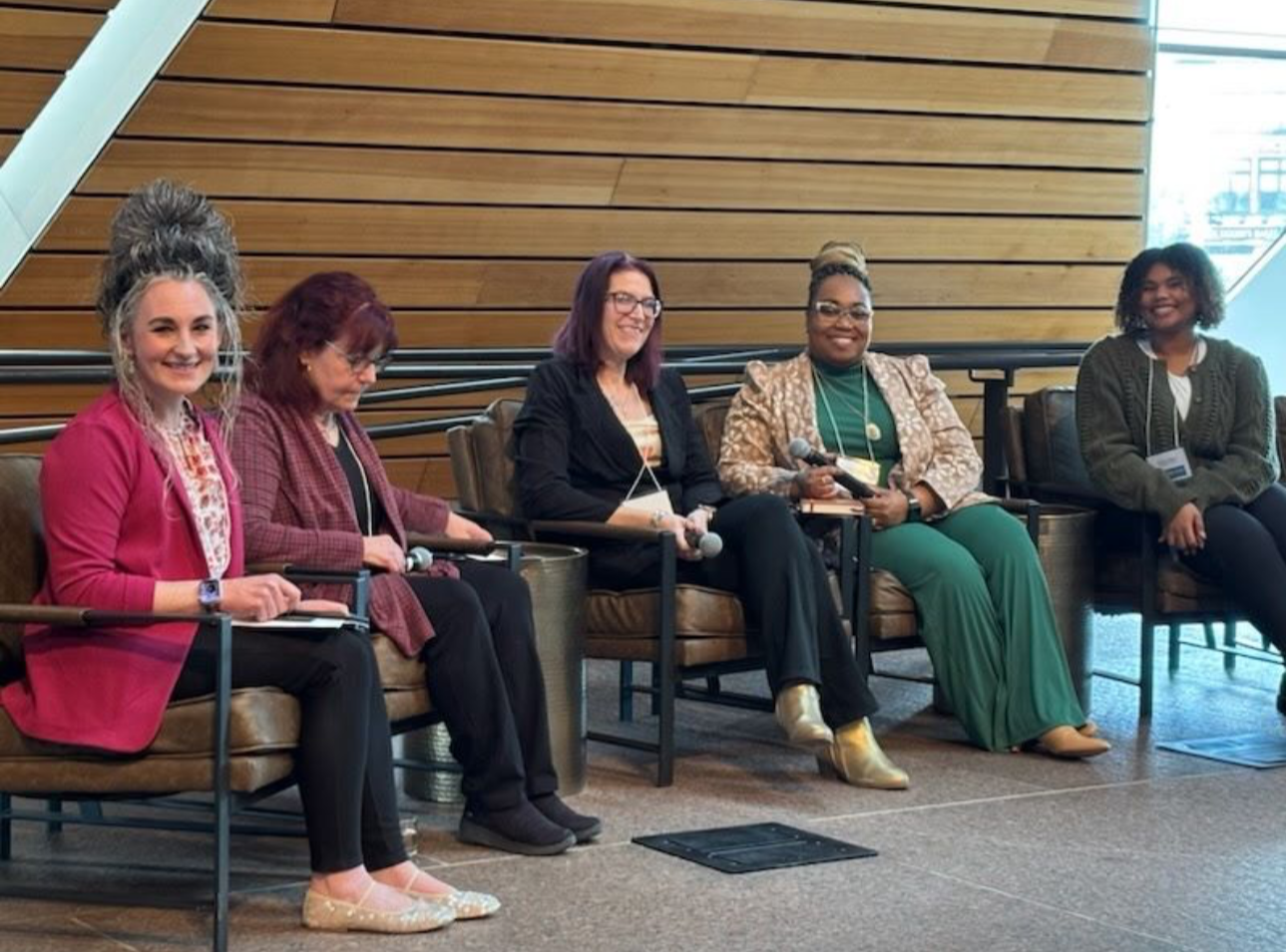 Five women sitting on a panel at a conference, smiling, holding microphones and notes, in a modern venue with wood-paneled wall behind them.