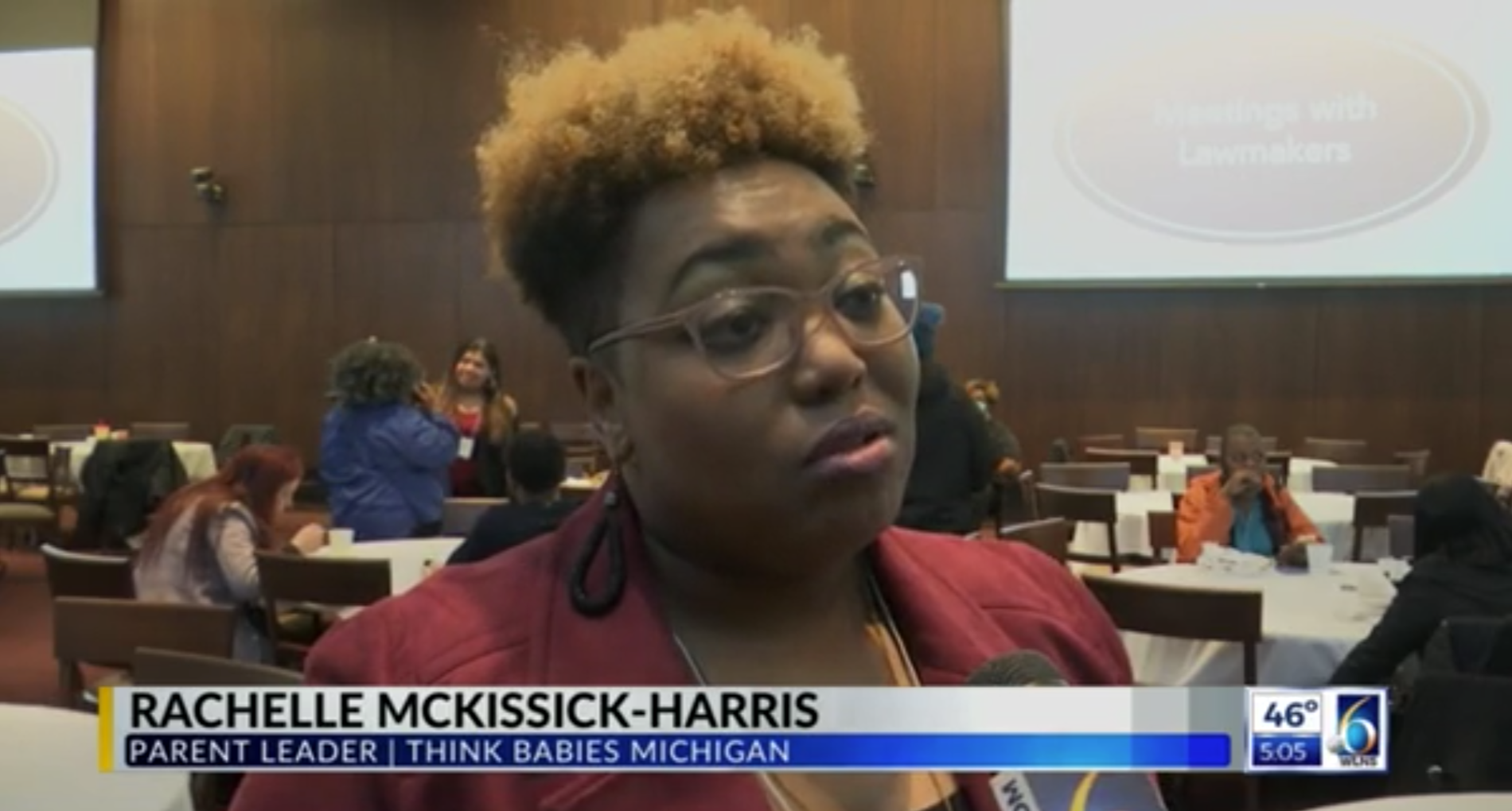 A woman being interviewed at a conference, with a name tag that reads 'RACHELLE McKISSICK-HARRIS'. In the background, there are other attendees sitting at tables in a conference room, some of whom are engaged in conversations.