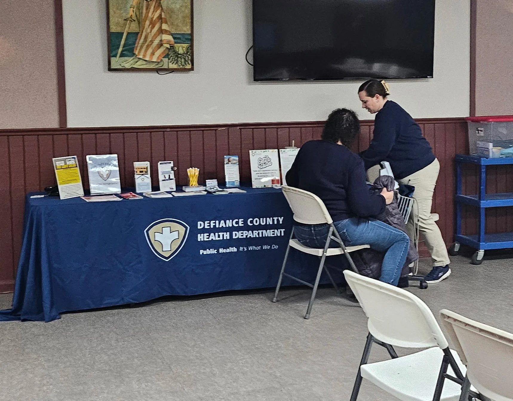 A table with a blue tablecloth displaying the logo and name of Defiance County Health Department, with informational brochures and supplies. Two women, one seated and one standing, are engaged in conversation in a room with gray carpet and wood-paneled walls. There are empty white chairs and a rolling cart nearby.