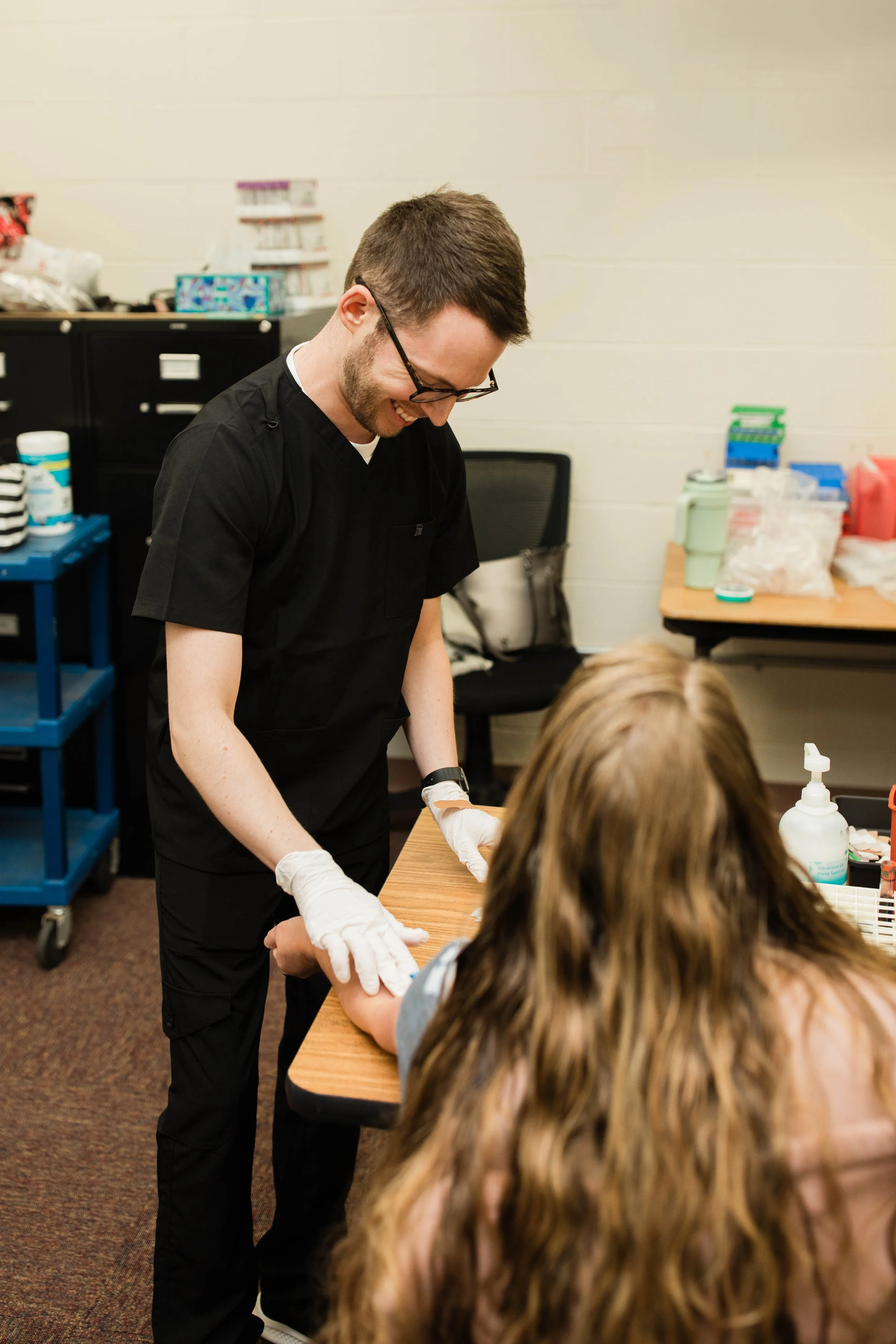 A healthcare professional with glasses and a beard, wearing black scrubs and white gloves, examining a patient's arm in a clinic or medical setting.