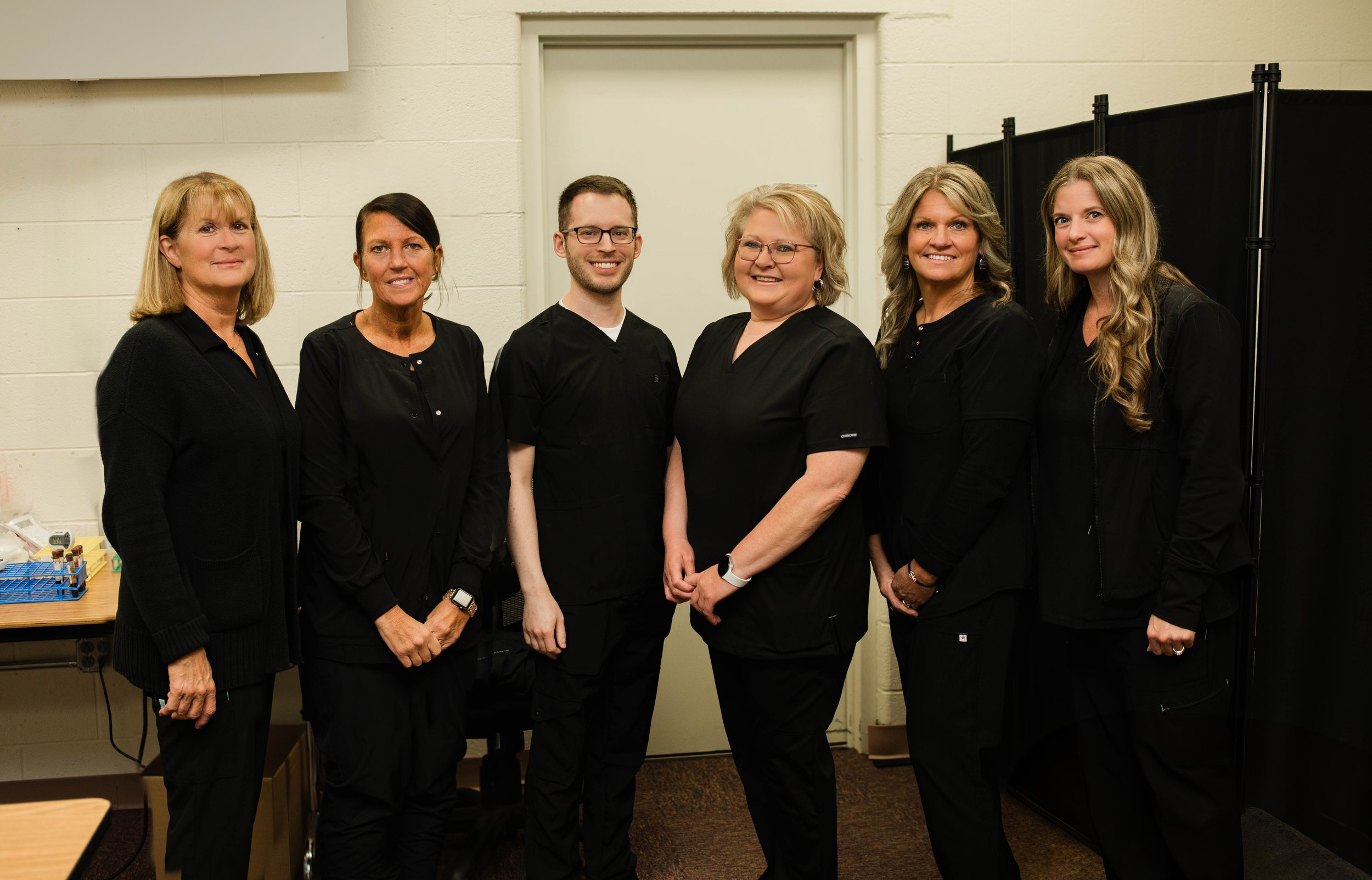 Group of six people, five women and one man, standing indoors in front of a white wall and black privacy screen, dressed in black, smiling at the camera.