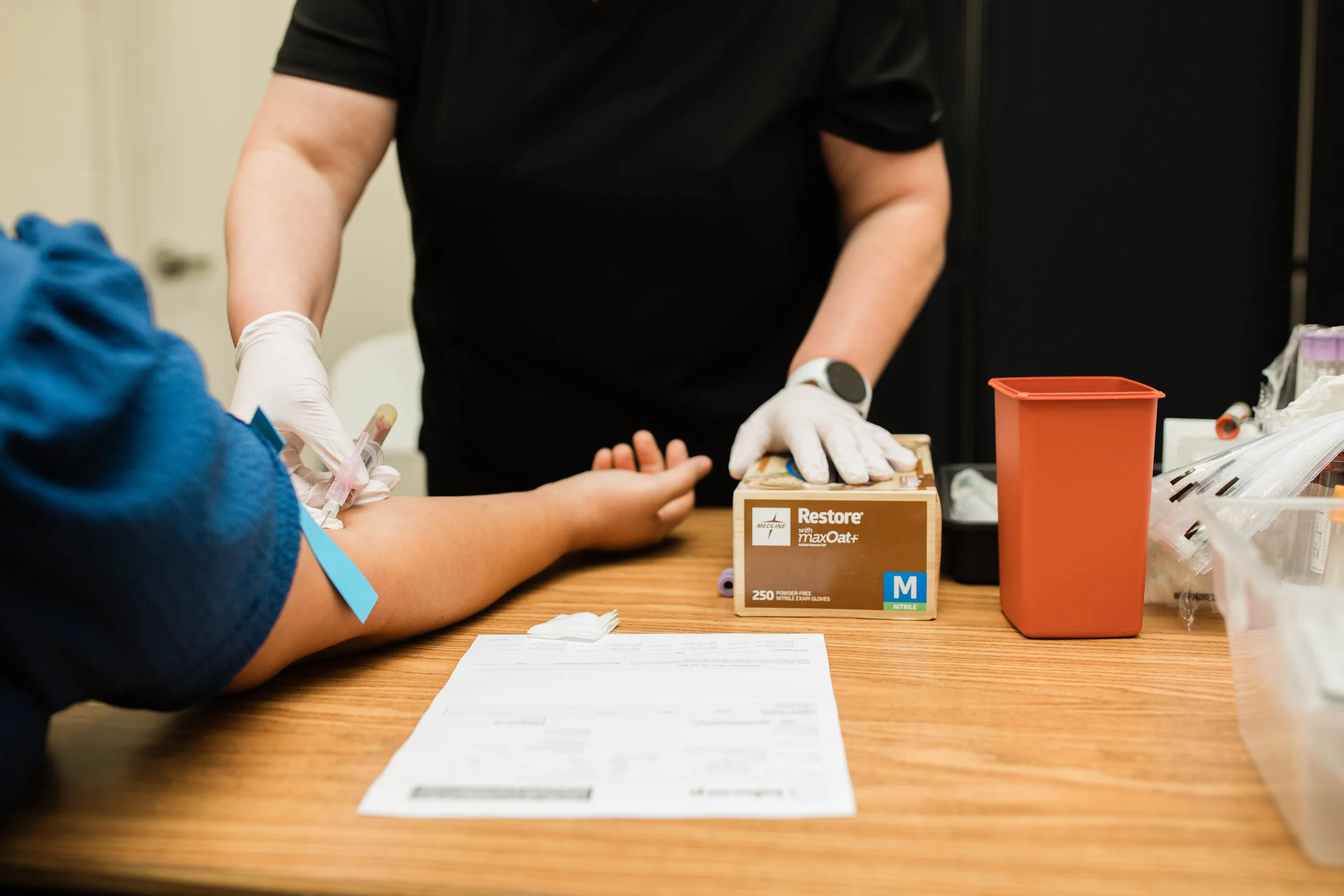 A person in black shirt and white gloves drawing blood from a patient for a medical test at a table with medical supplies and paper work.