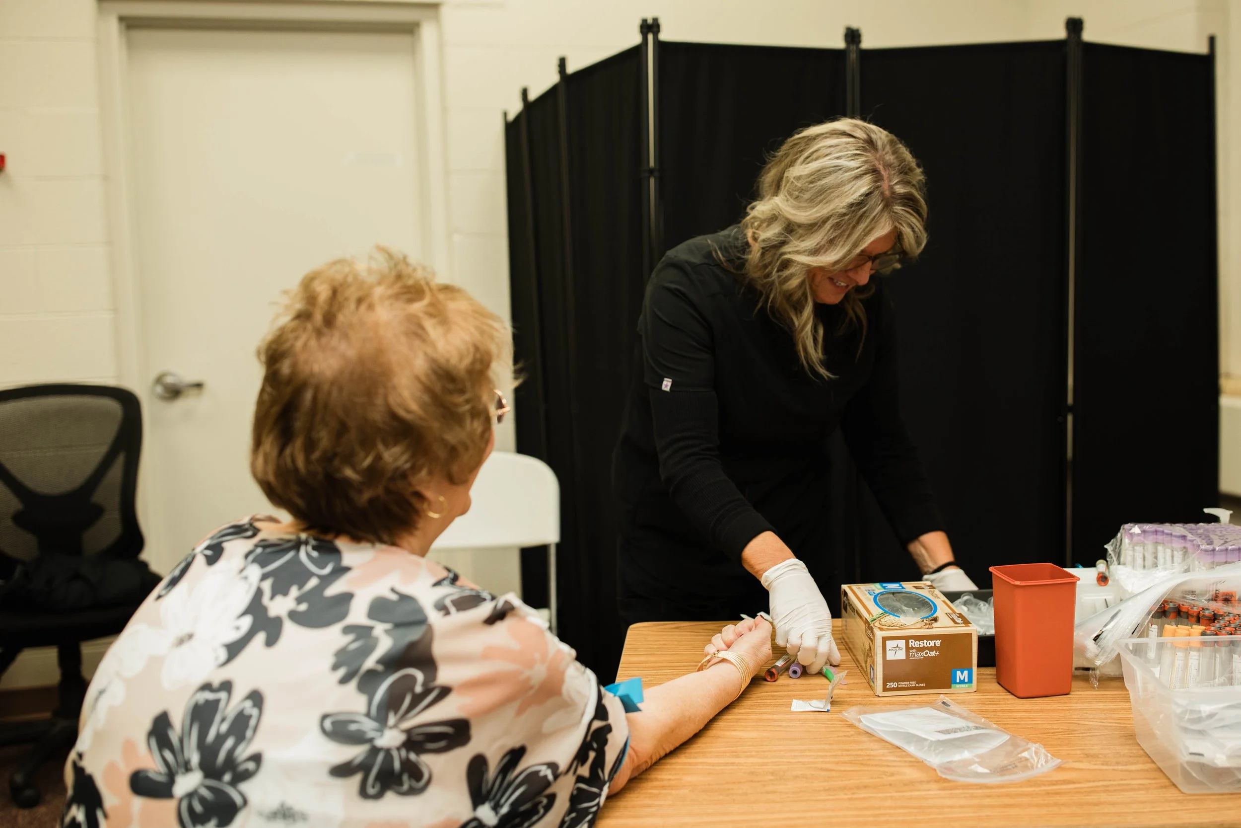 Two women, one with gray hair and glasses and the other with curly hair, are engaged in an activity at a table in an indoor setting. The woman with gray hair is sitting and the other woman is standing, both involved in what appears to be a crafting or medical activity with various supplies on the table. There is a black folding screen and a white door in the background.