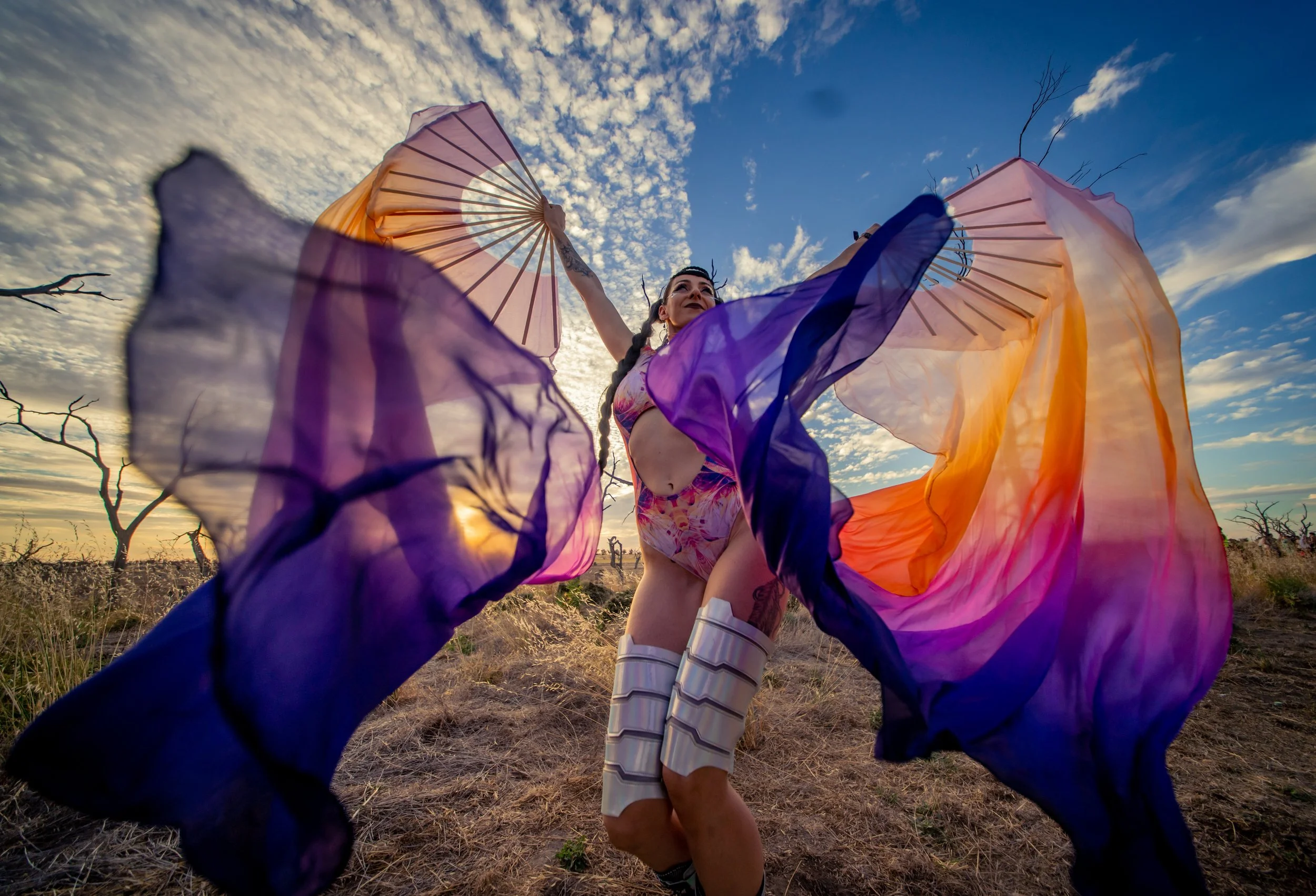 A woman standing outdoors in a dry field during sunset or sunrise, holding a large, colorful fabric umbrella while lifting her arm, with the sky filled with scattered clouds in the background.