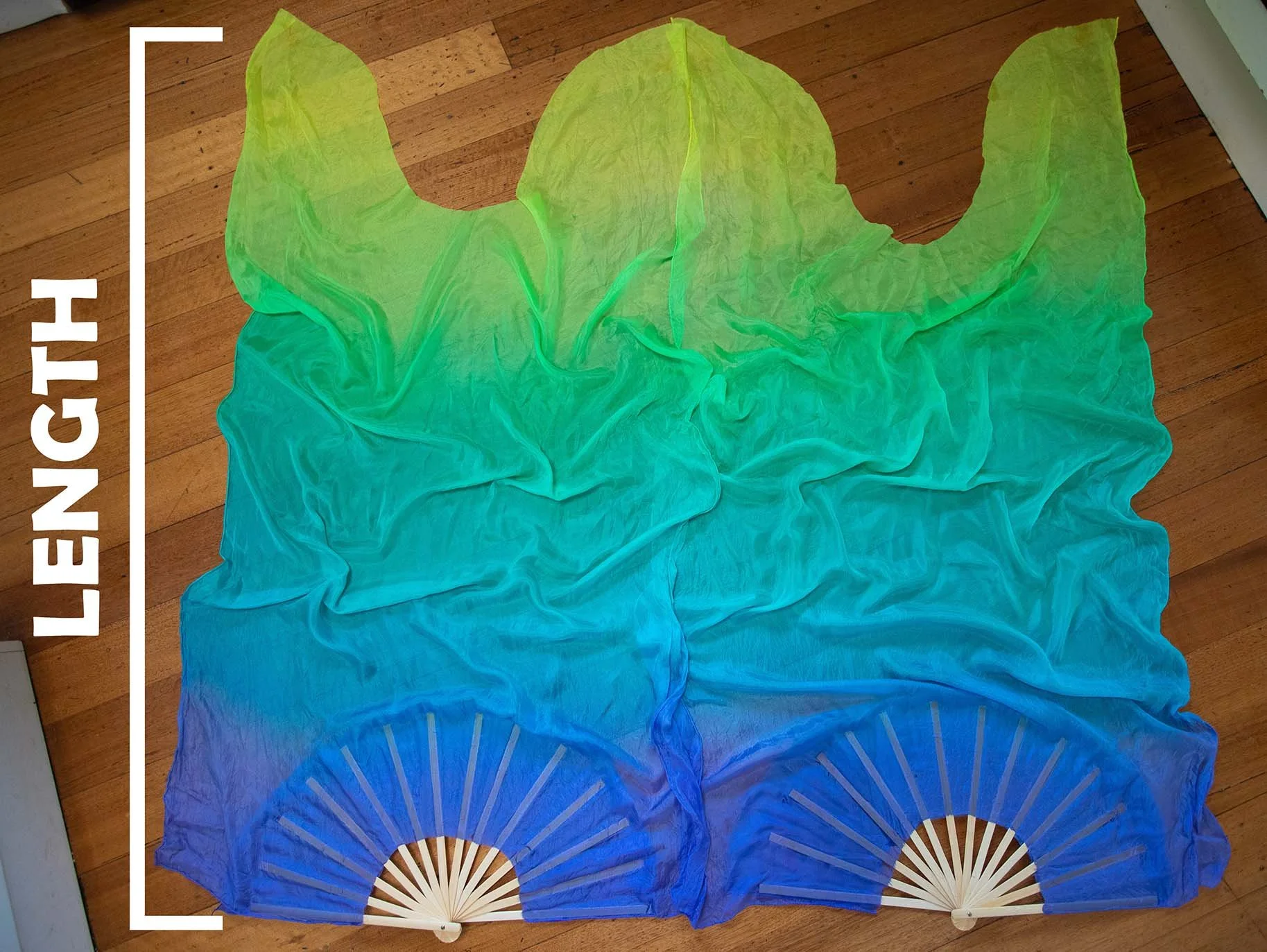 Three hand fans with colorful fabric in shades of green, blue, and purple, spread out on a hardwood floor. The word 'LETTERING' is written vertically along the left side of the image.