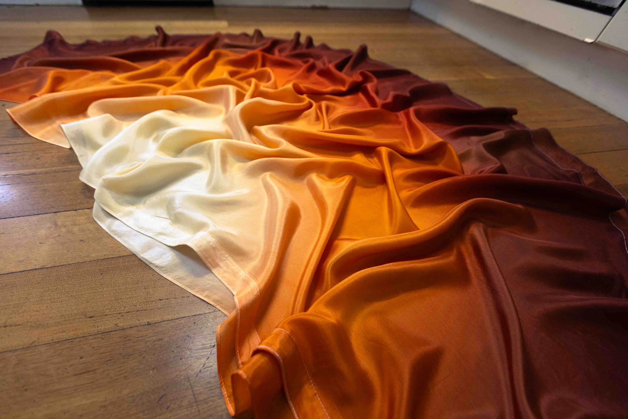 A fabric scarf with an ombre gradient from white to yellow, orange, and dark brown, laid out on a wooden floor.
