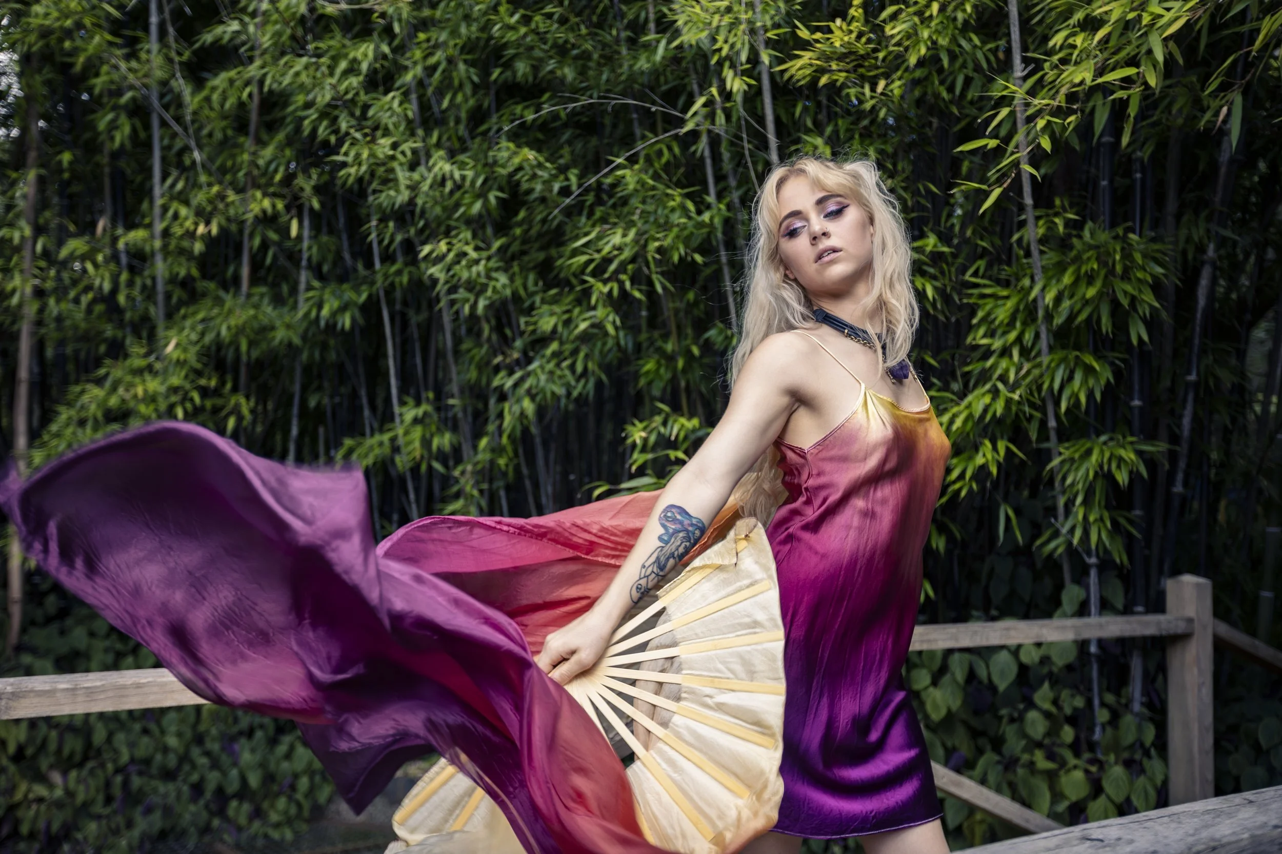A woman with blonde curly hair, tattoos on her arm, wearing a colorful dress, poses outdoors against a green leafy background, holding an open folding fan with a purple veil flowing around her.