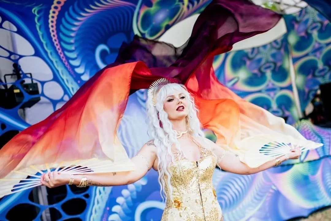 A woman with long white hair dressed in a gold and white outfit, holding fans and wearing a crown, stands against a vibrant, psychedelic background with flowing colorful fabrics.