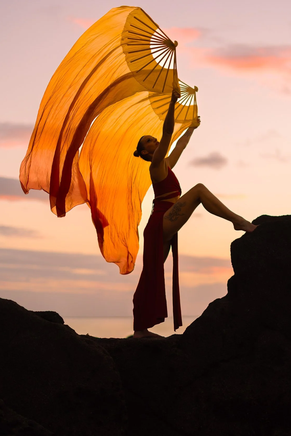 Woman standing on rocks at sunset, holding large yellow fans, wearing red dress with a cutout, with the sky and water in the background.