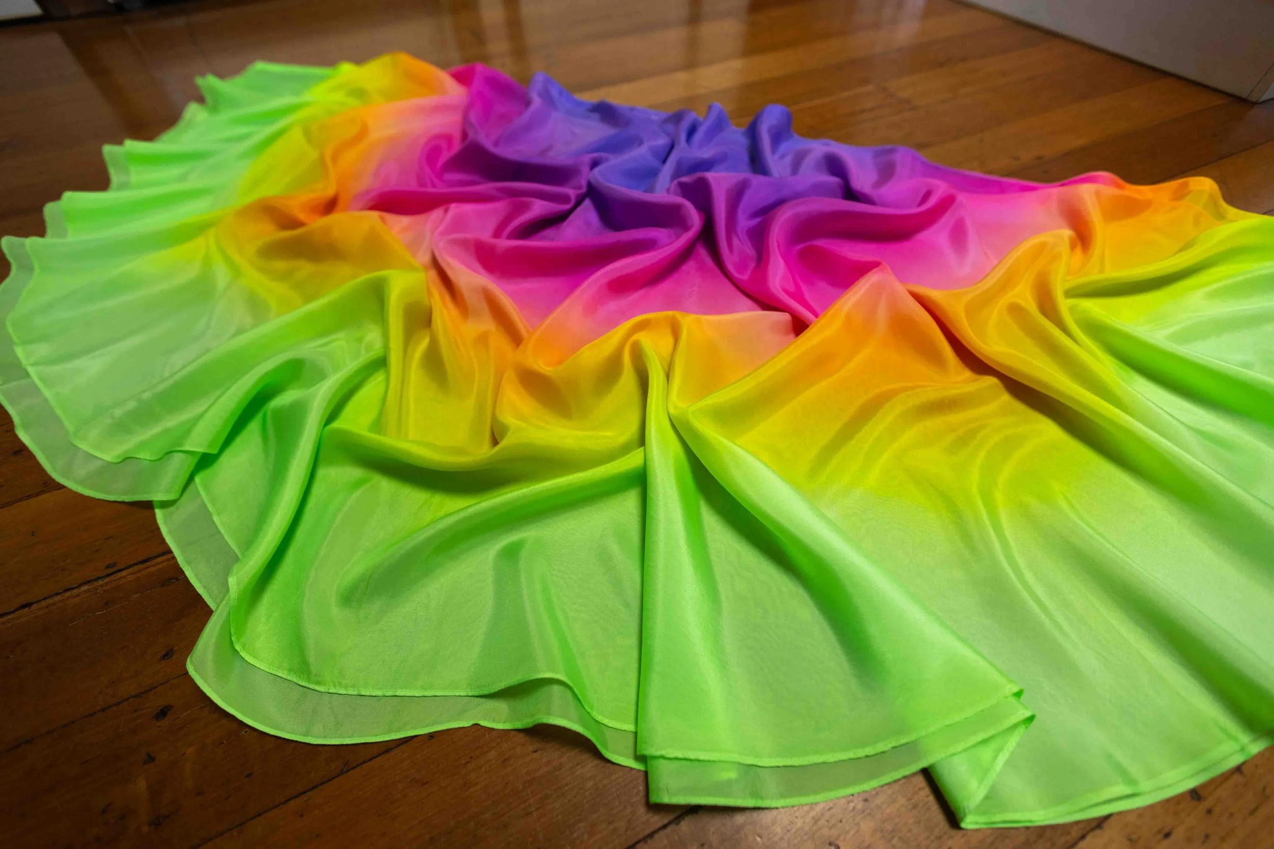 A colorful rainbow ruffled fabric with neon yellow, orange, pink, purple, and blue sections, laid flat on a wooden floor.