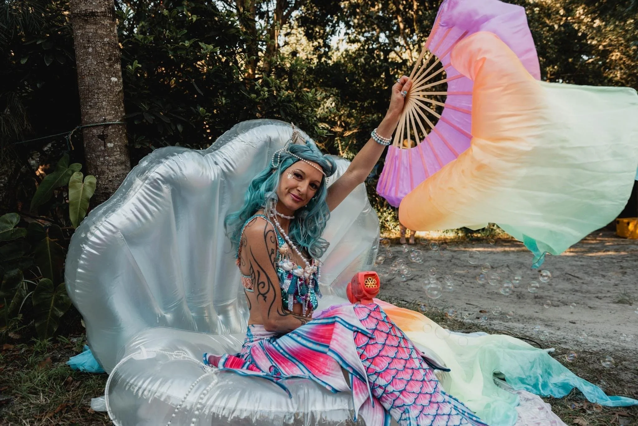 A woman with blue hair and tattoos, dressed in fantasy-style clothing, holding a large pastel-colored parasol outdoors among trees, surrounded by bubbles.