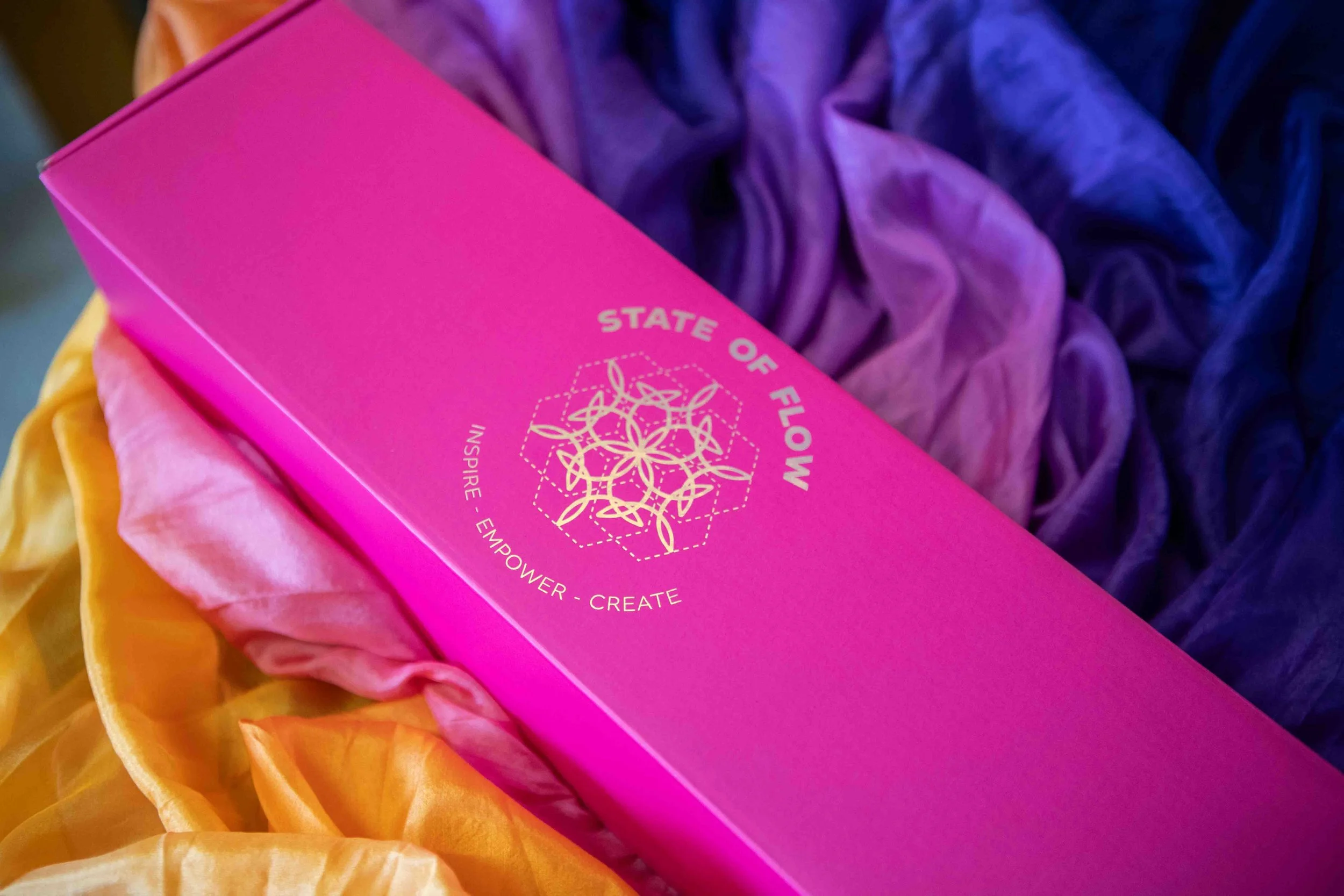 Close-up of a pink box with gold foil details, resting on multicolored fabric. The box features a geometric floral design and the text 'STATE OF FLOW' with the words 'Empower - Create - Inspire' below.