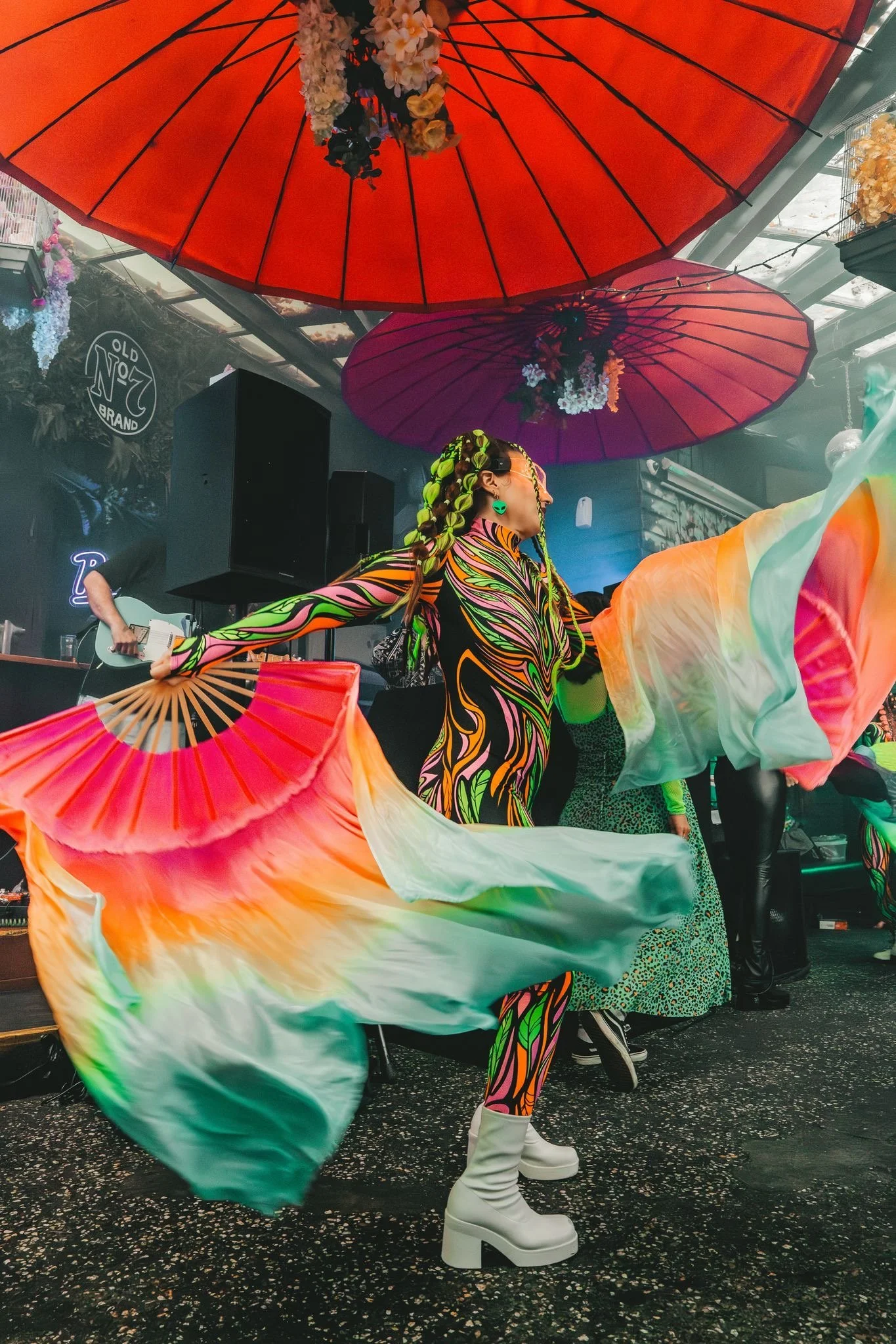 A performer dressed in colorful, psychedelic-patterned clothing with long braids and white platform boots is dancing with rainbow-colored flowing scarves under large orange and purple umbrellas decorated with flowers, in a lively indoor setting.