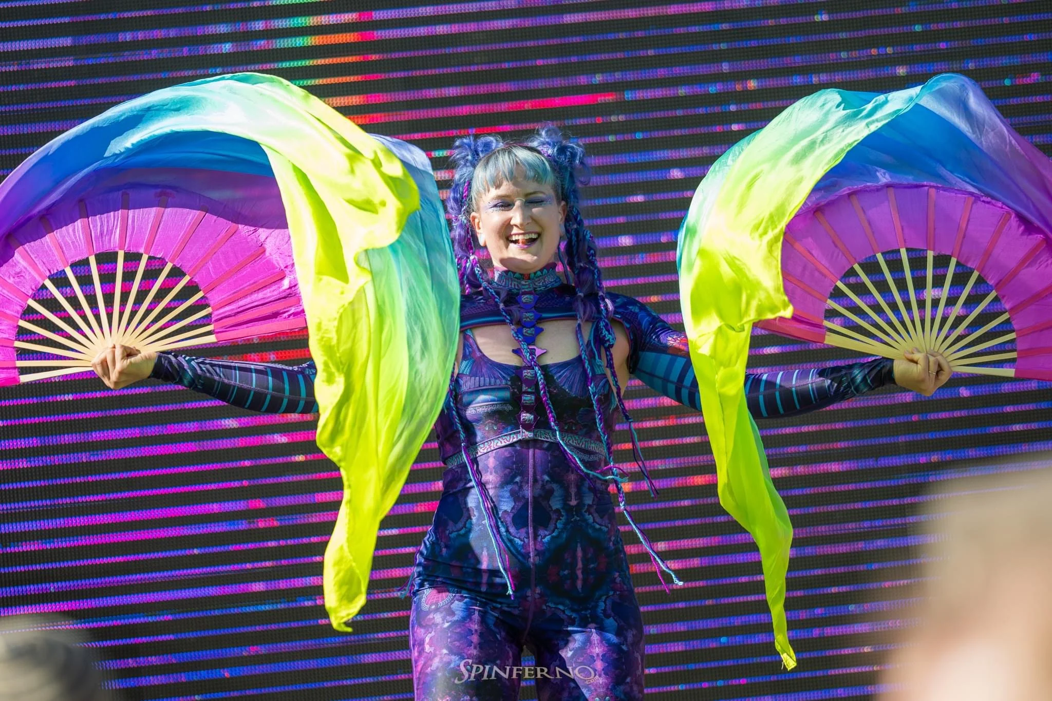 Performer with colorful hair and rainbow attire holding two rainbow fans on stage, smiling in front of a large digital screen display.