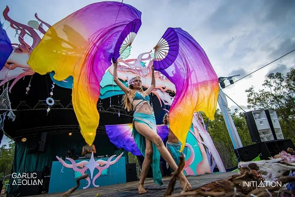 A woman performing a dance with large purple, yellow, and pink fabric fans at an outdoor festival stage with trees and a cloudy sky in the background.
