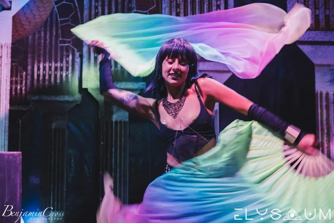 A woman with black hair, wearing a black top and jewelry, is dancing with colorful fabric in a dimly lit setting.