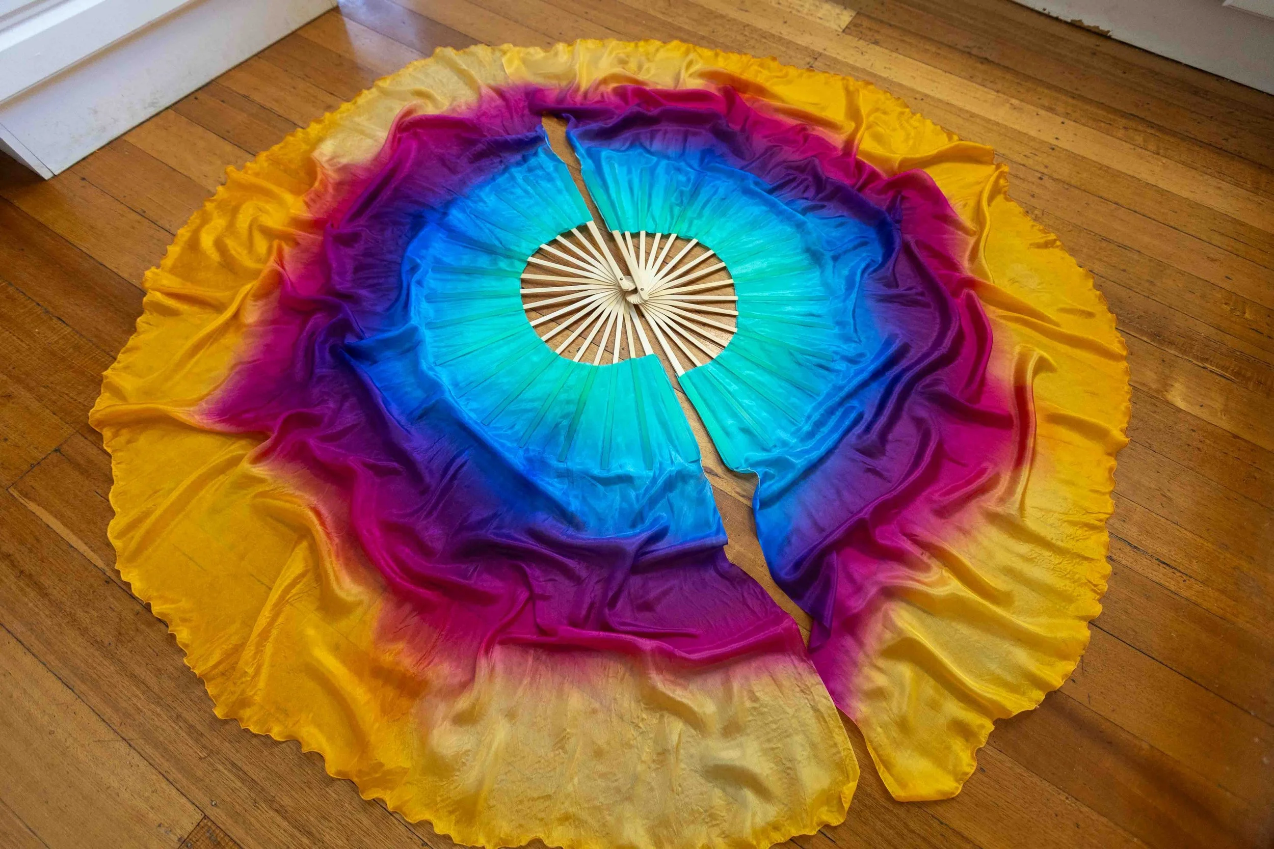 Colorful fabric arranged on a wooden floor in a circular pattern with rainbow colors, centered around a small round wooden object.