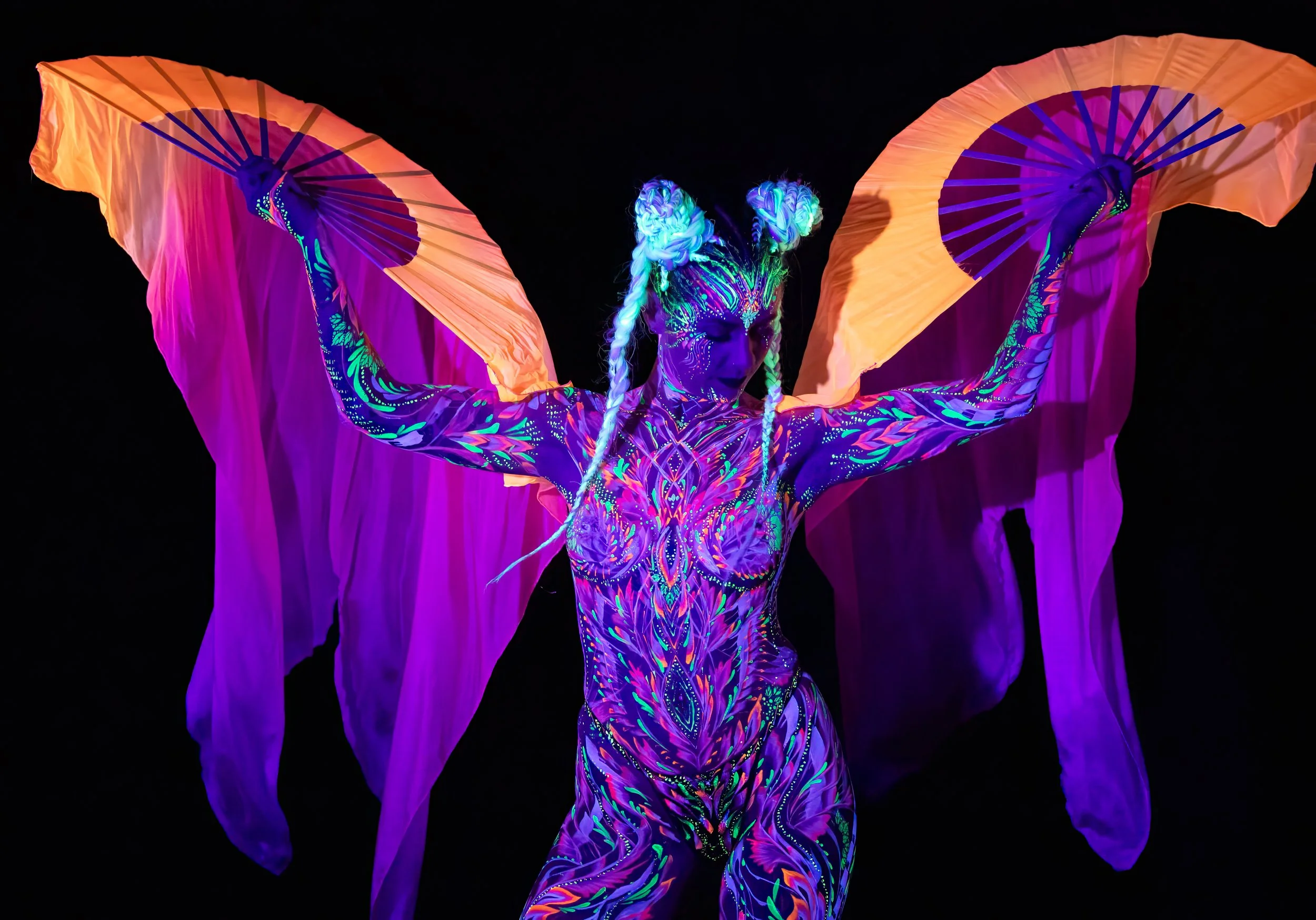 Performer with neon body paint and flowing orange fabric holding large orange fans in a dark setting.