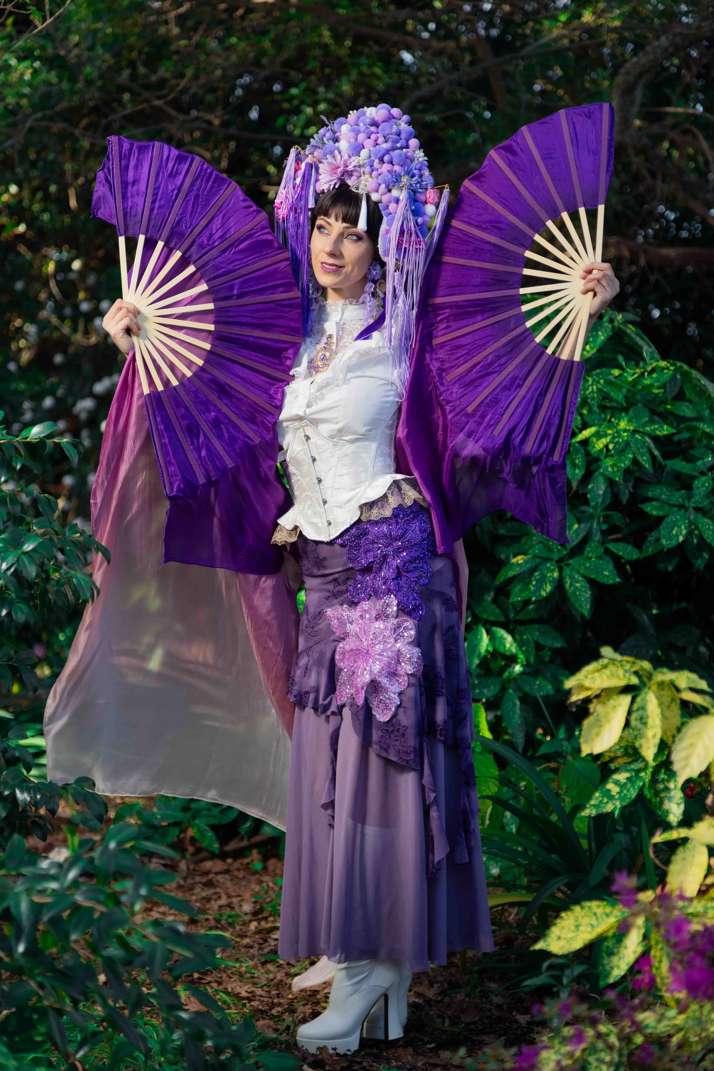 Woman dressed in elaborate purple and violet costume with large purple fans and floral headpiece, standing in a lush green forest.