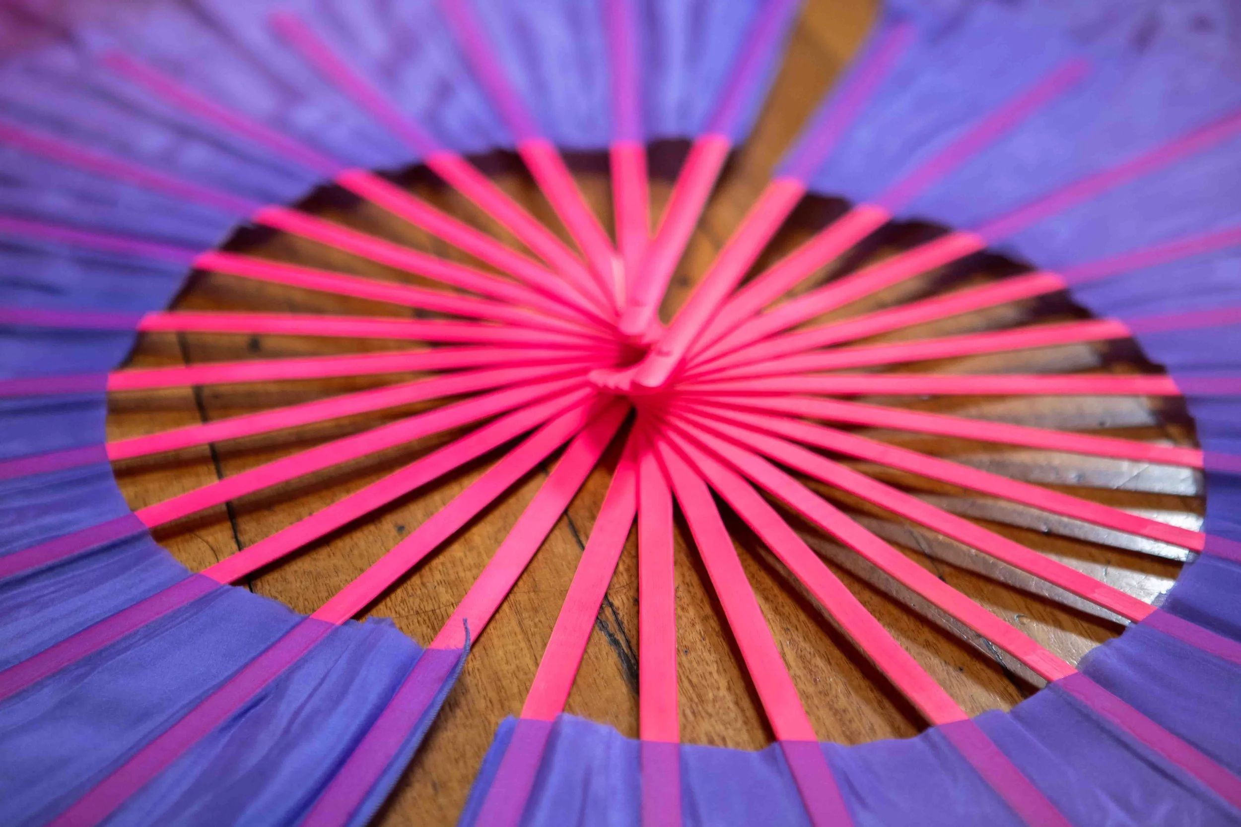 Close-up photo of a colorful paper parasol with pink and blue spokes on a wooden surface.