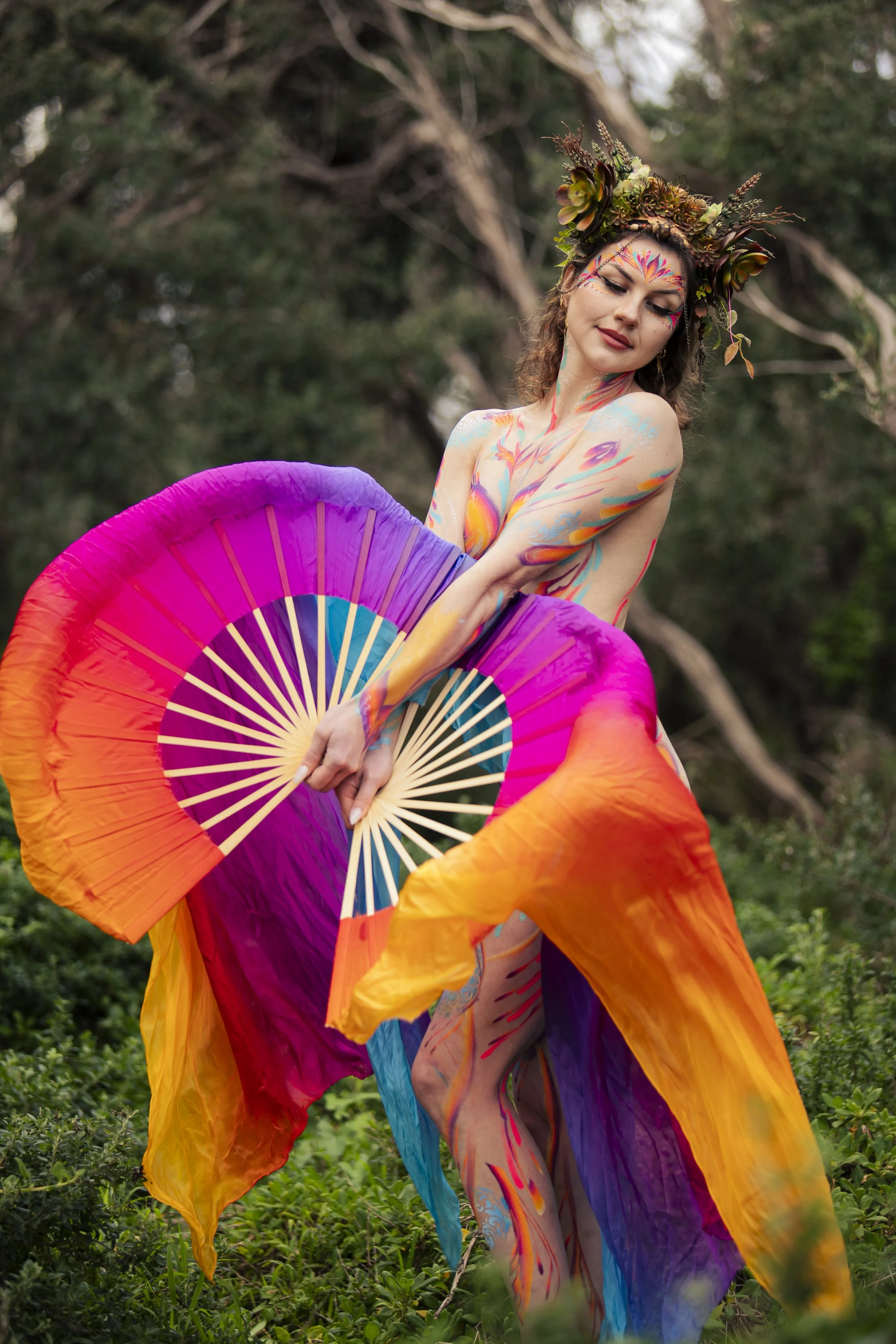 A woman with body paint and a leafy crown is holding a multicolored fan in an outdoor setting with greenery and trees in the background.