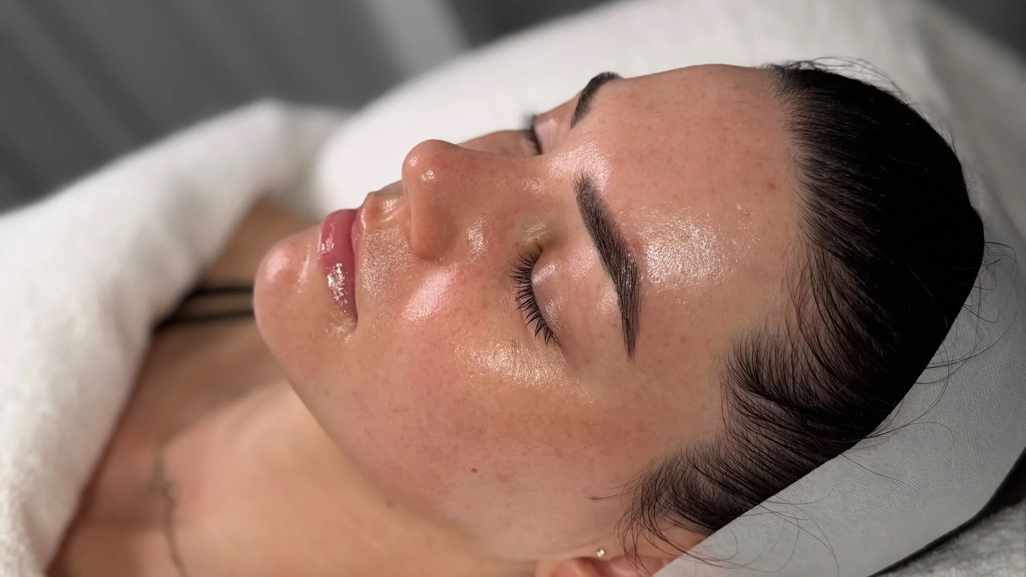 Person receiving a facial cleansing treatment, lying down with eyes closed, using cotton pads to apply skincare, in a spa or skincare clinic.