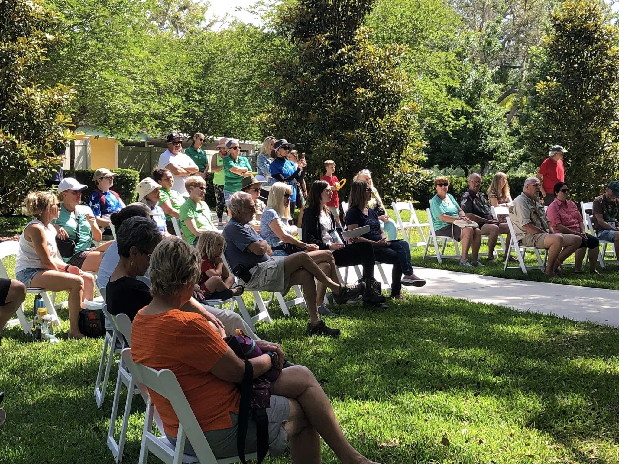 A group of people sitting on white chairs outdoors on green grass, attending an event under large trees with green leaves.