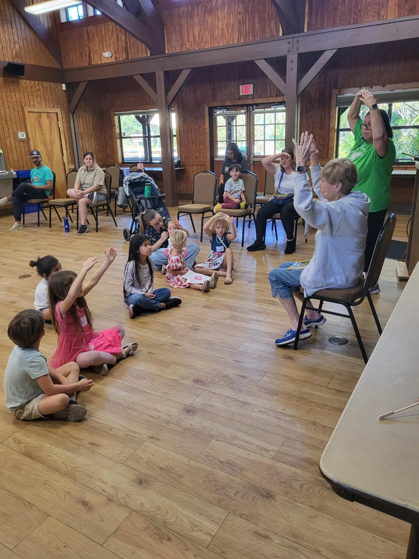 A group of children sitting on the floor raising their hands, while adults sit and listen to a woman with glasses and short hair who is engaging with the children in a room with wooden walls and large windows.