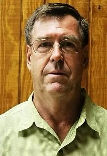 A middle-aged man with glasses and short, dark hair standing in front of a wooden background.