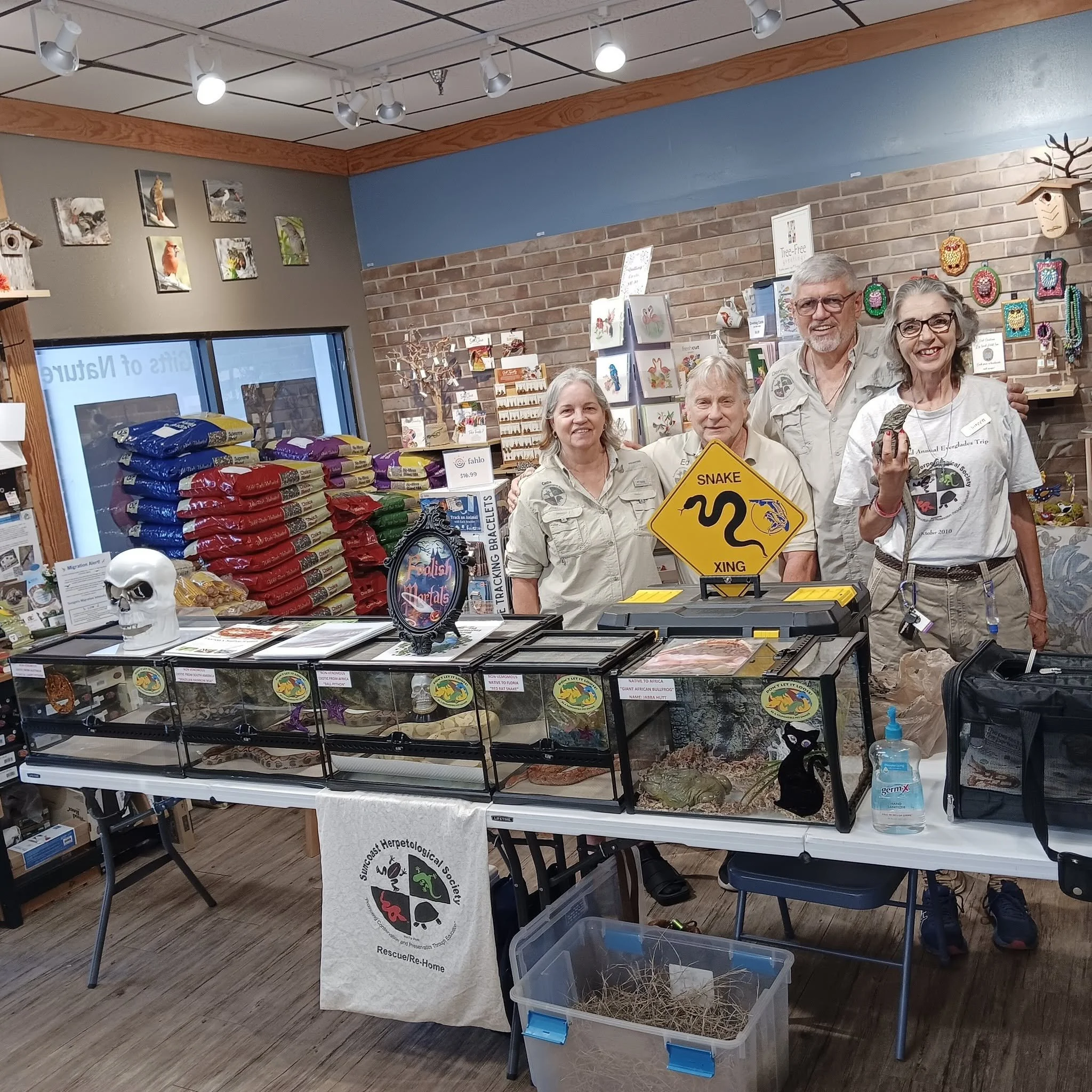 A group of people standing behind a table at an indoor event, with various animal-related displays, posters, and supplies around them, including a snake crossing warning sign on the table.