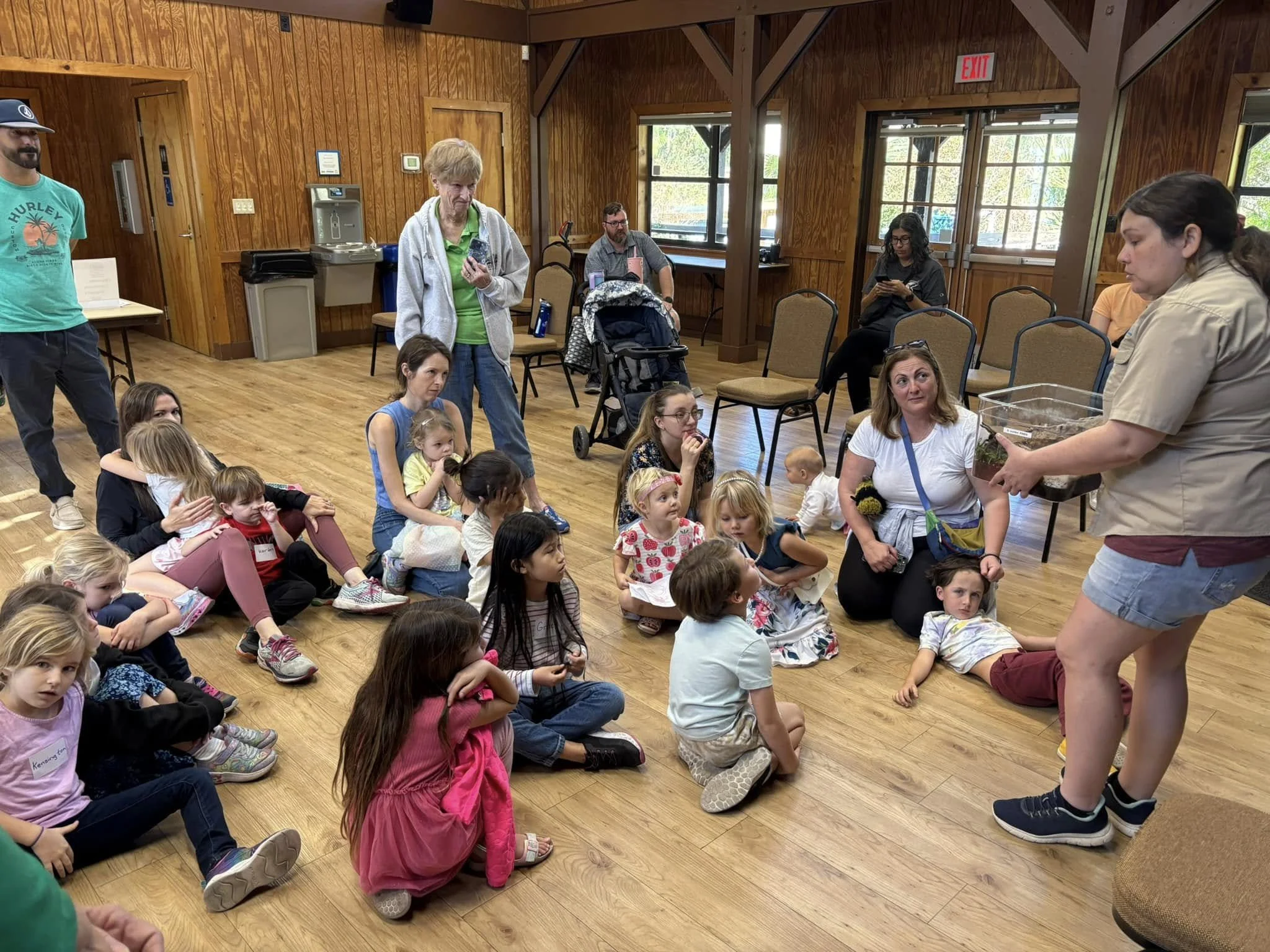 A group of children and adults sitting and standing in a wooden-finished room, with some looking at a woman in uniform holding a glass container with insects. Some people are using their phones, and natural light is coming in through windows.