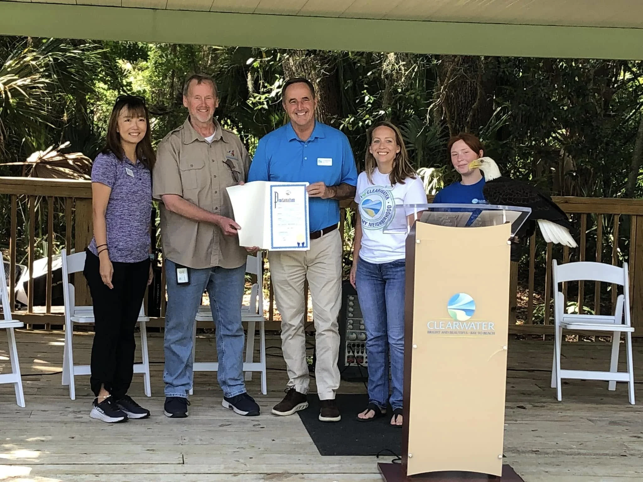 Five people standing together outdoors, smiling, with a man holding a certificate. An eagle is perched nearby, and a sign reads 'Clearwater Beach and Natural Habitat'.