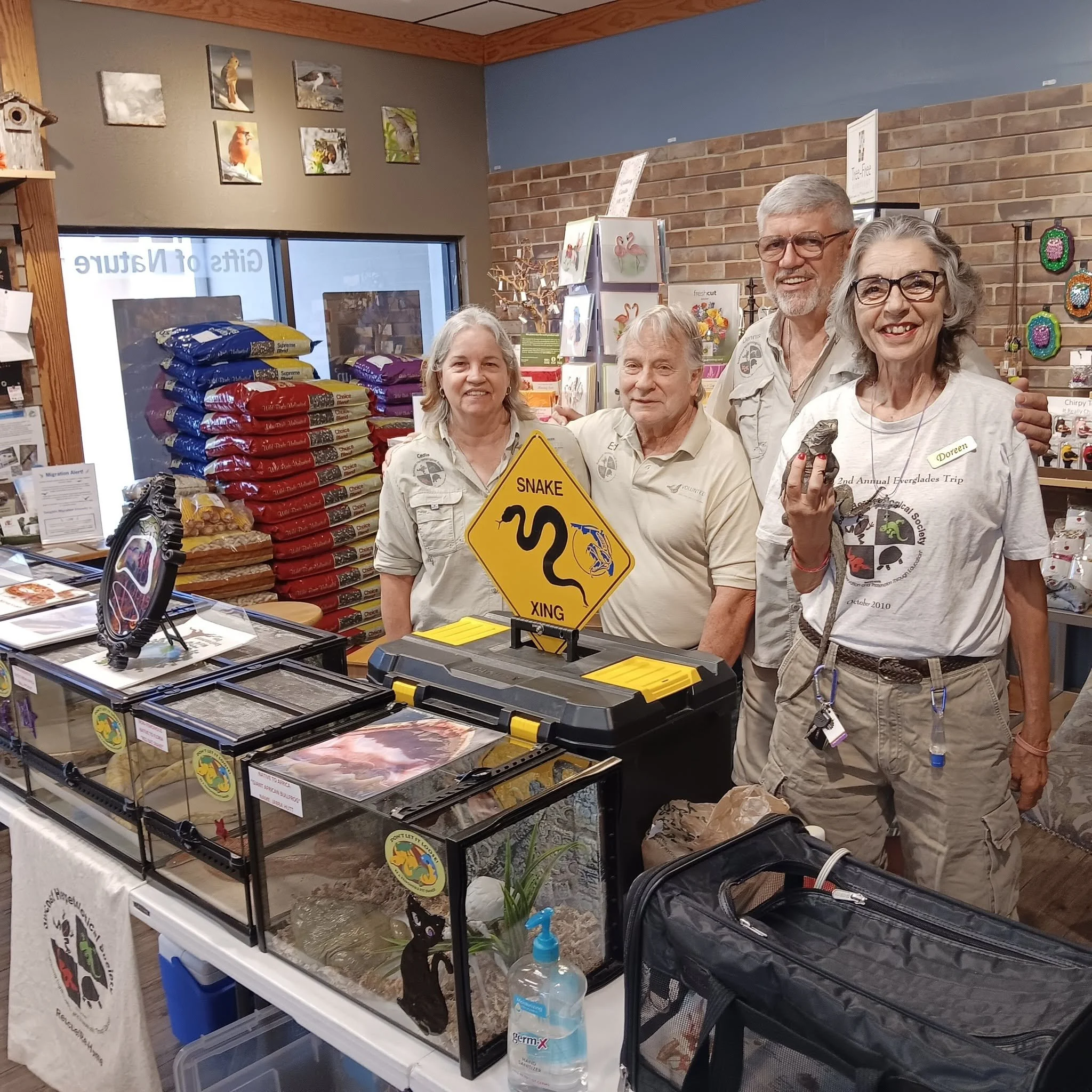 Group of five elderly people standing inside a store near pet supplies and clothing, smiling at the camera. Stacked bags of pet food on a table, colorful wall art, brick wall, and a large window are visible in the background.