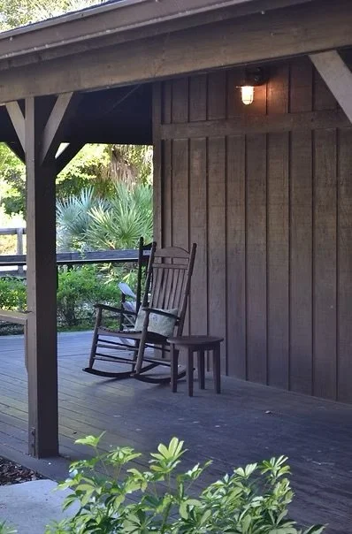 A wooden porch with a rocking chair and a small stool, with greenery in the background and a small light on the wall.