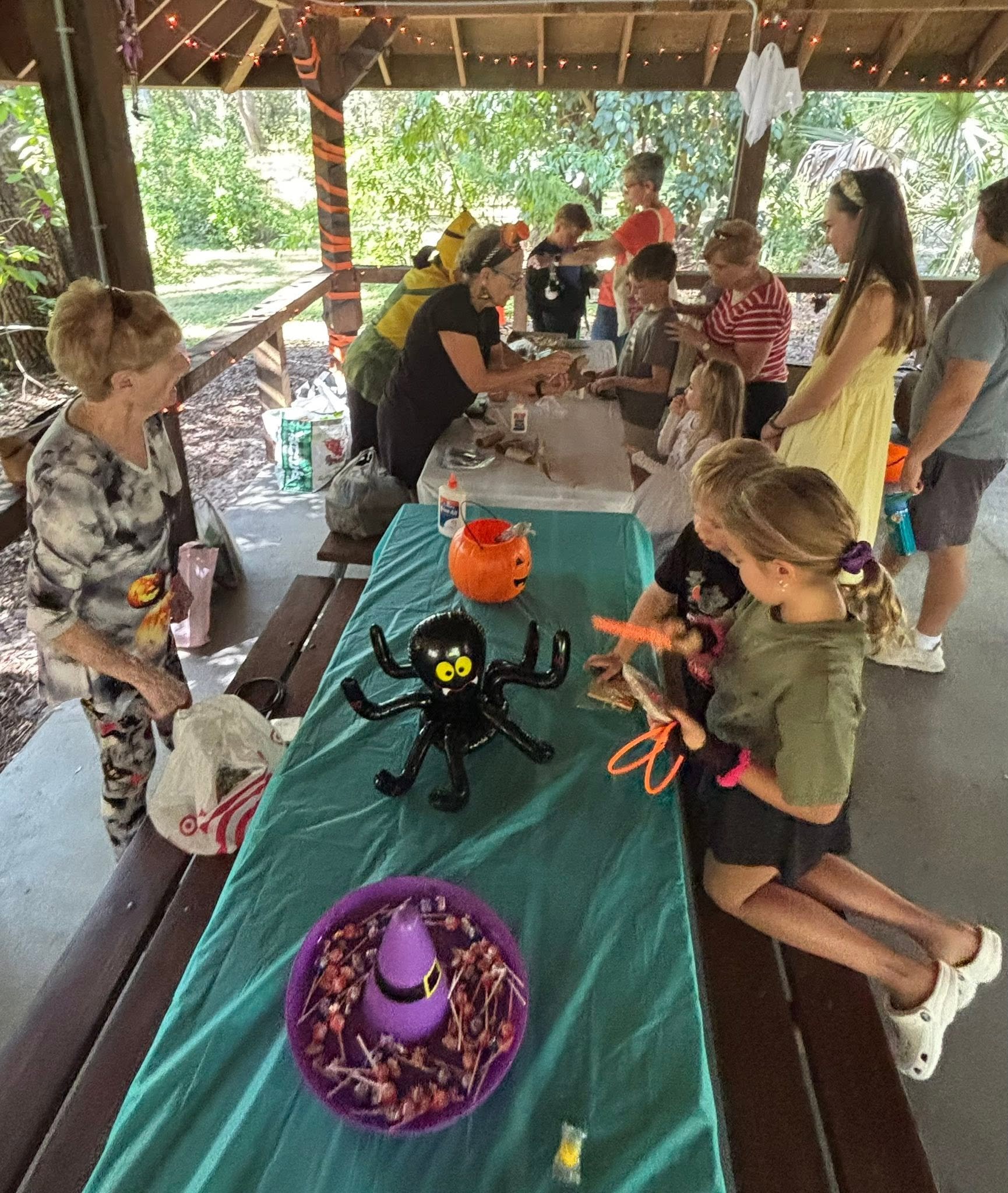Children and adults participating in a Halloween-themed craft or activity under a wooden shelter with string lights, decorated with Halloween decorations including a black spider with yellow eyes, orange pumpkin, and witch hat, on a green tablecloth.