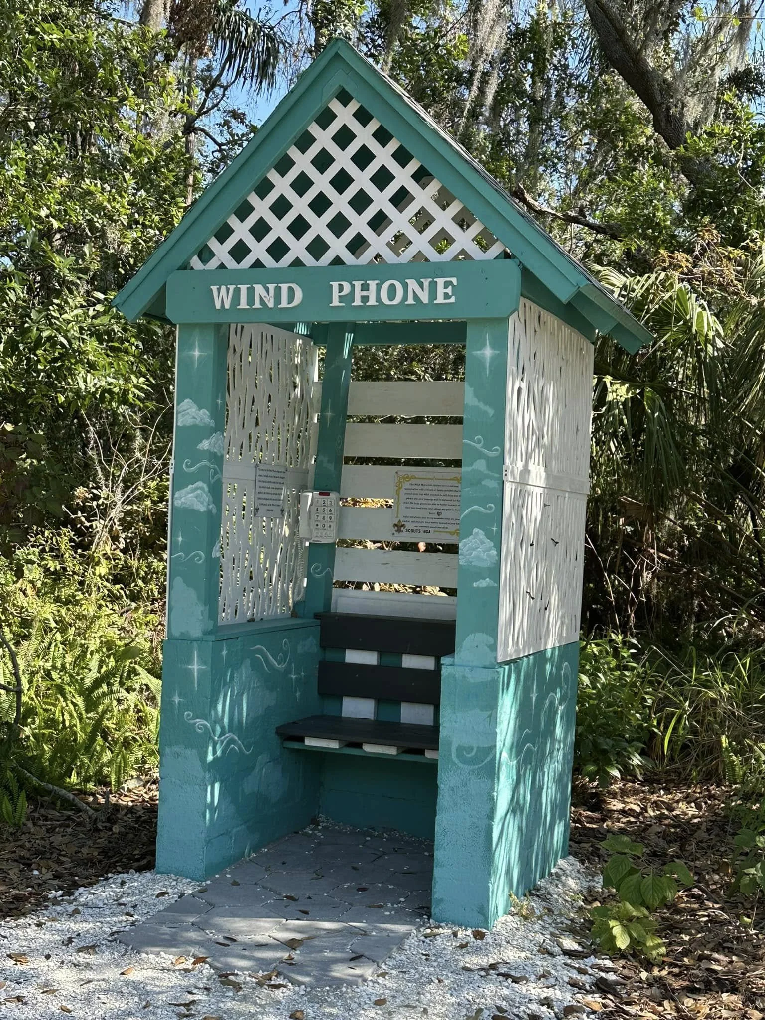 A teal-colored outdoor telephone booth labeled 'WIND PHONE' with decorative clouds and mountain paintings, surrounded by greenery and trees.