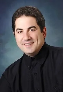 Portrait of a man with dark hair wearing a black shirt, smiling against a blue background.
