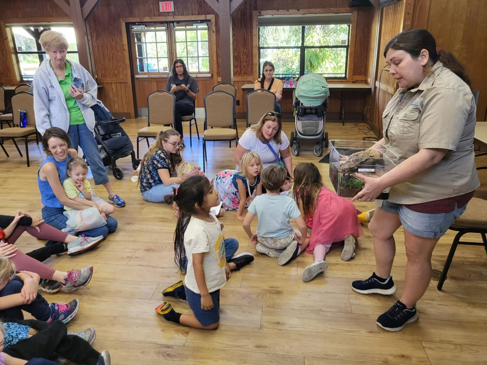 A group of children sitting on the wooden floor of a room, listening to a woman who is holding a terrarium with a large reptile inside. Several adults are sitting or standing nearby, observing the scene.