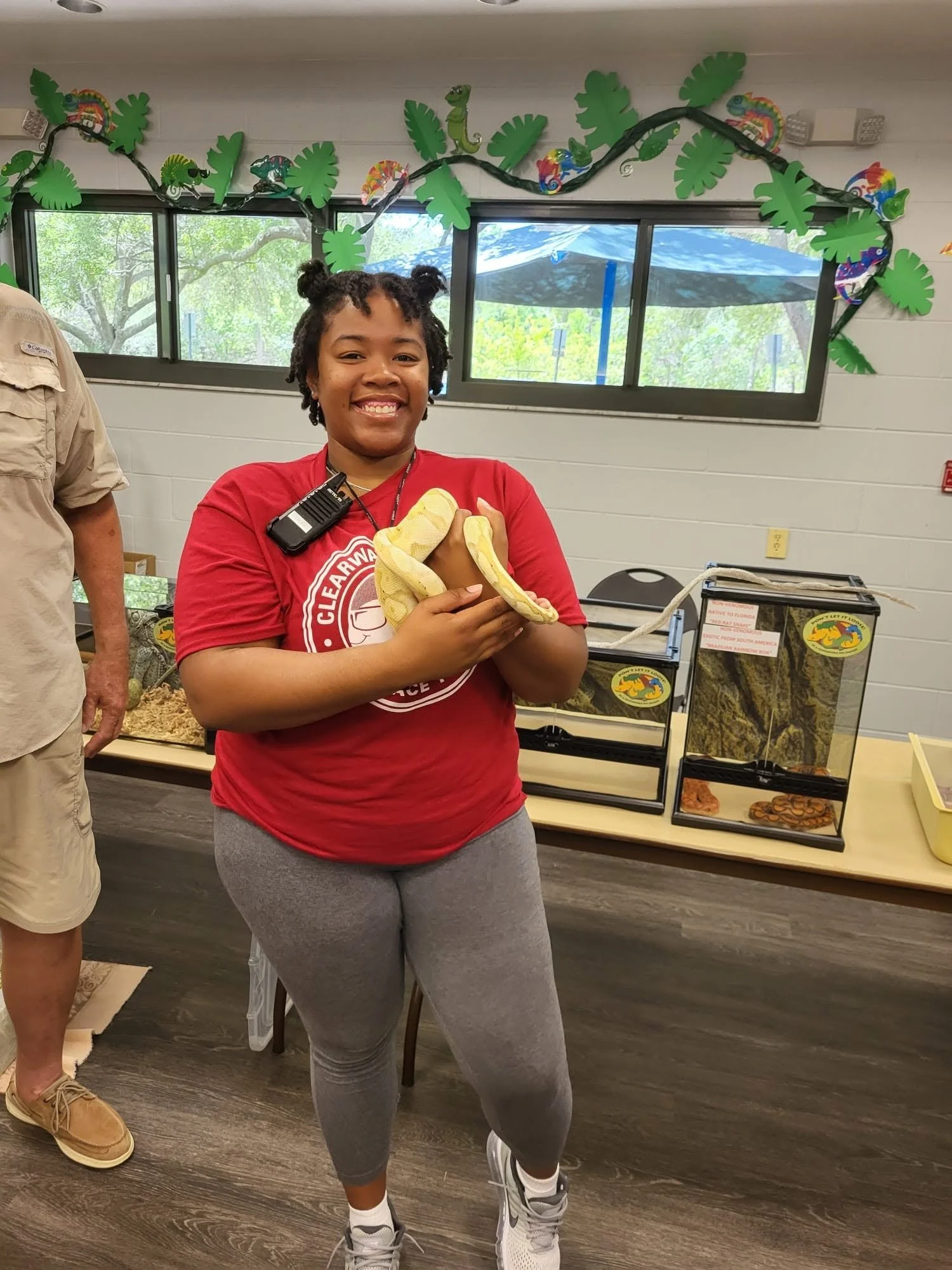 A young woman with two small buns in her hair, wearing a red t-shirt with a white graphic and gray sweatpants, holding a yellow and white albino snake with a smiling expression. She is standing indoors in front of a decoration with green leaves and c