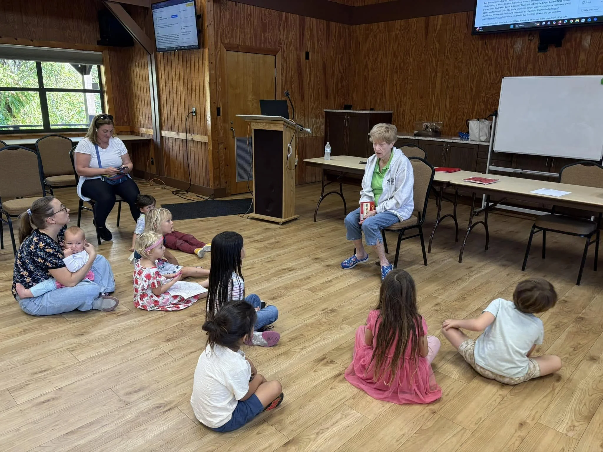 A woman with short blonde hair wearing a white jacket and green shirt sitting on a chair and holding a book, with children sitting cross-legged on the wooden floor in front of her. Several children and another woman with glasses are sitting nearby, l