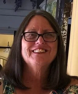 A smiling woman with long dark hair, glasses, and a patterned top, in an indoor setting with plants and a window in the background.